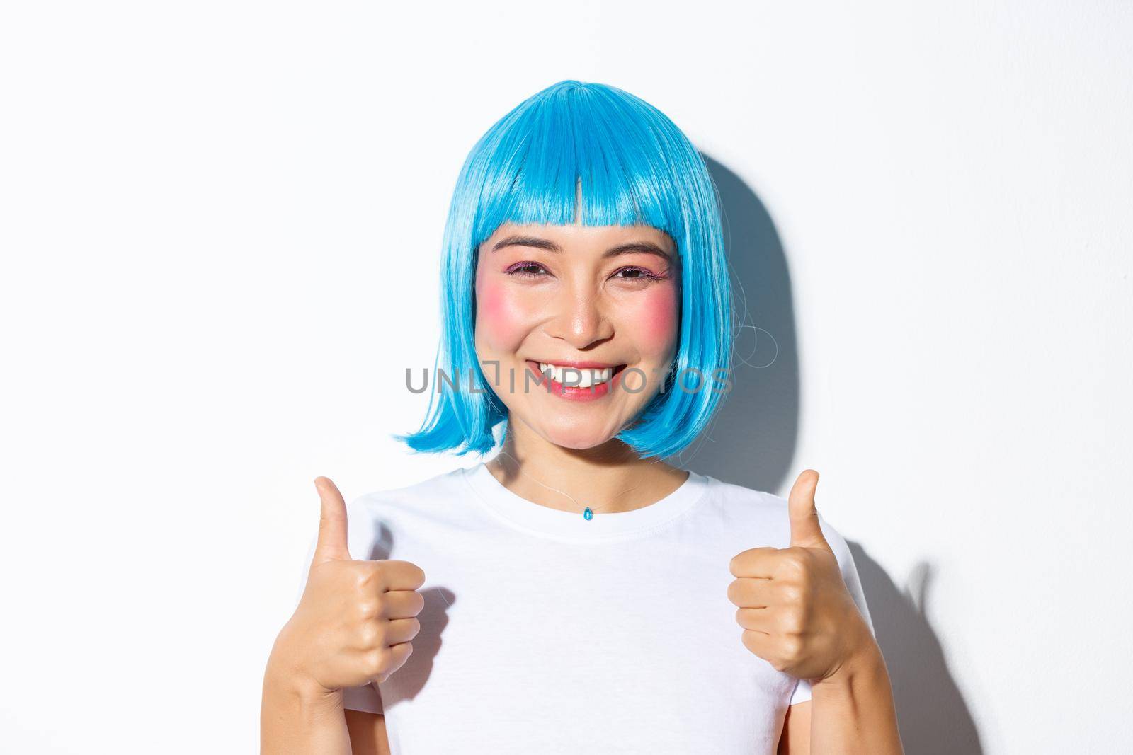 Close-up of happy asian girl in blue wig, smiling and showing thumbs-up in approval, like something good, standing over white background.
