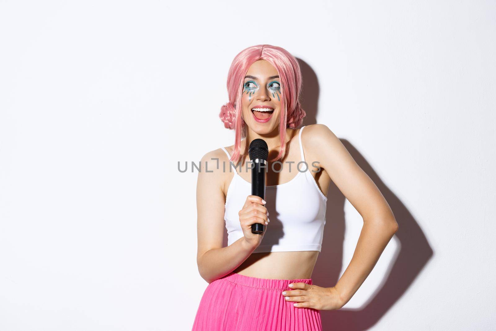 Image of beautiful smiling girl in pink wig, singing song in microphone, wearing halloween costume for party, standing over white background.