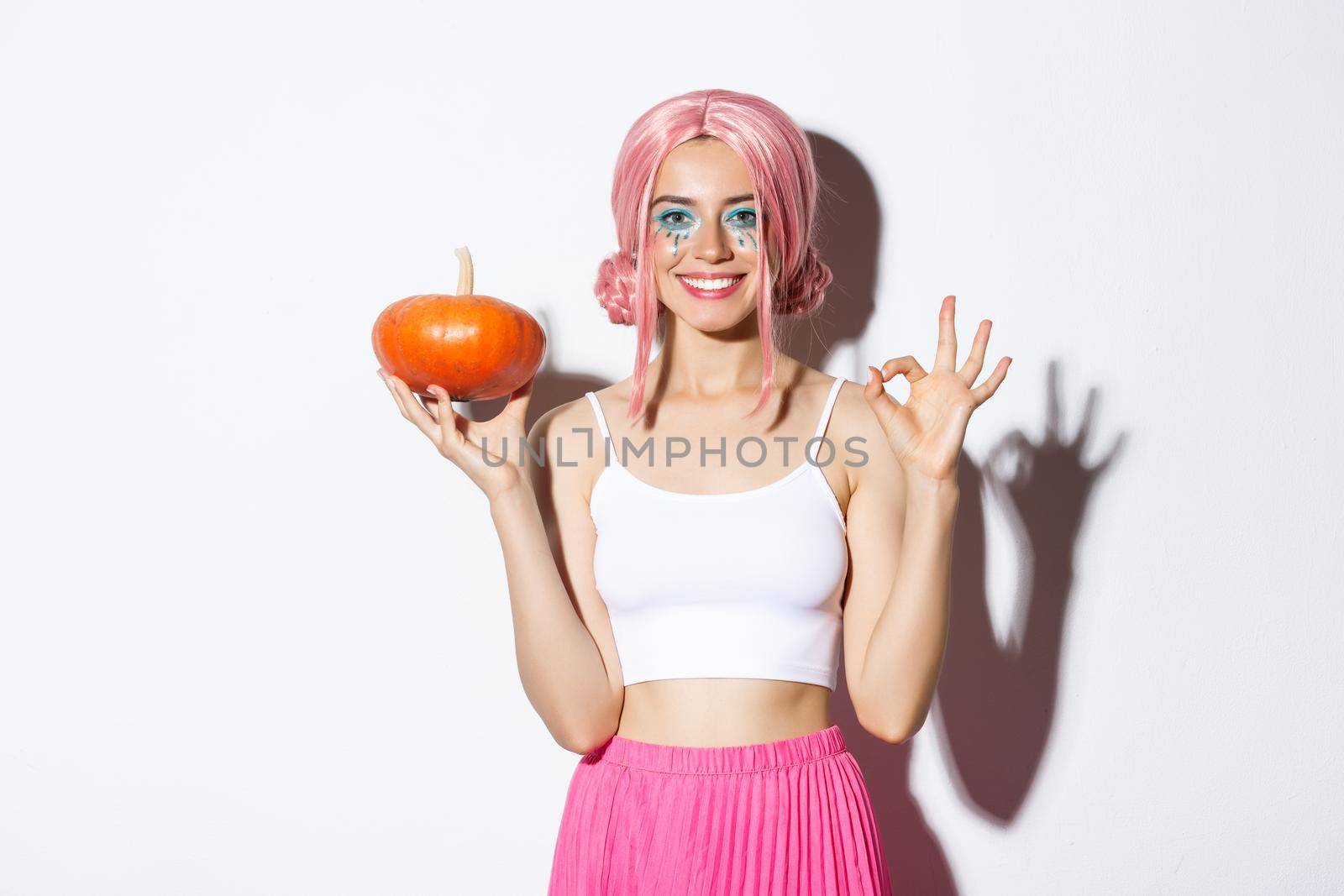 Portrait of happy girl celebrating halloween, showing small pumpkin and okay gesture, wearing cute pink wig.