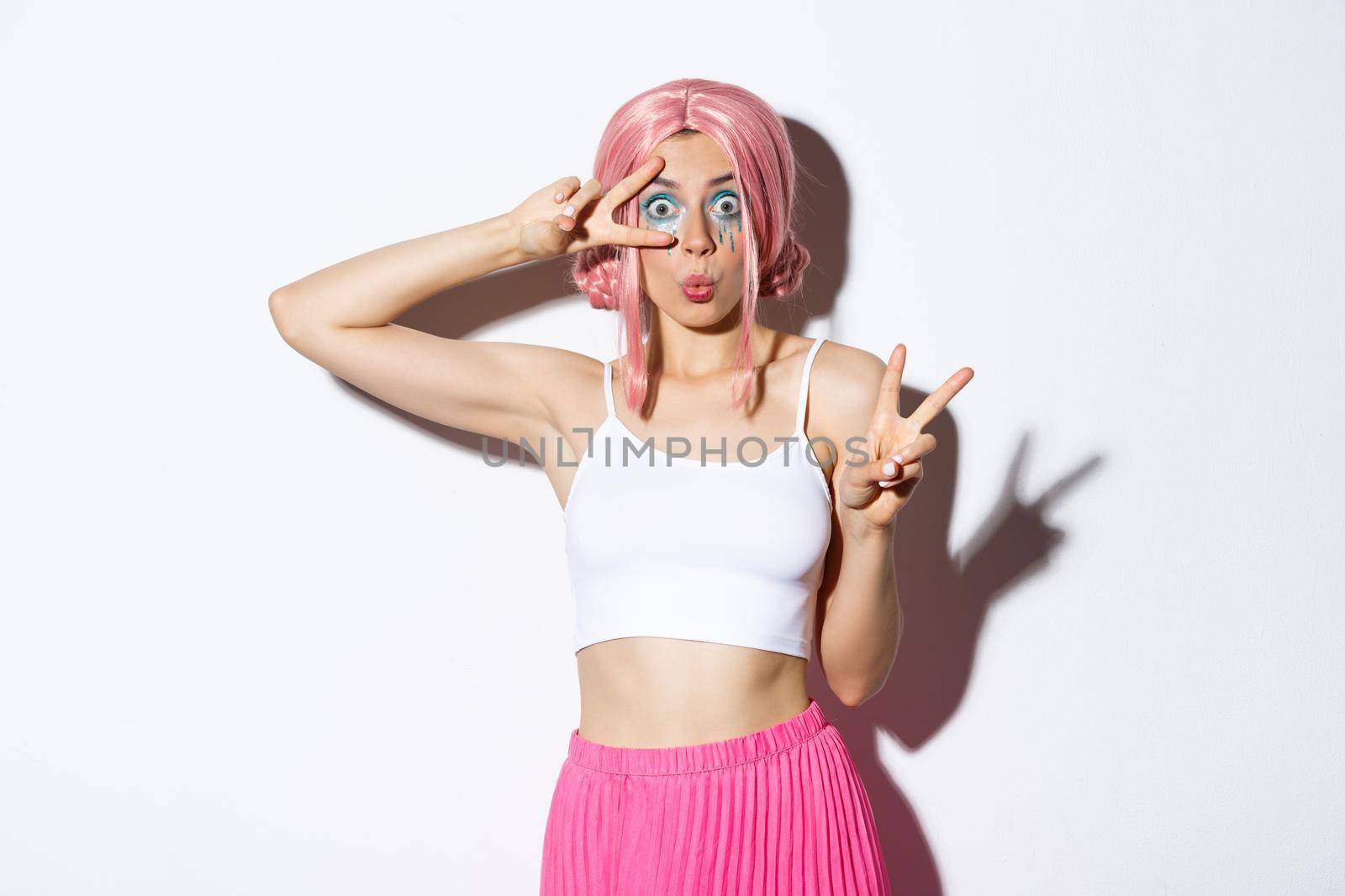 Image of amazed pretty girl with pink anime wig and colorful makeup, showing peace signs, celebrating halloween, standing over white background.