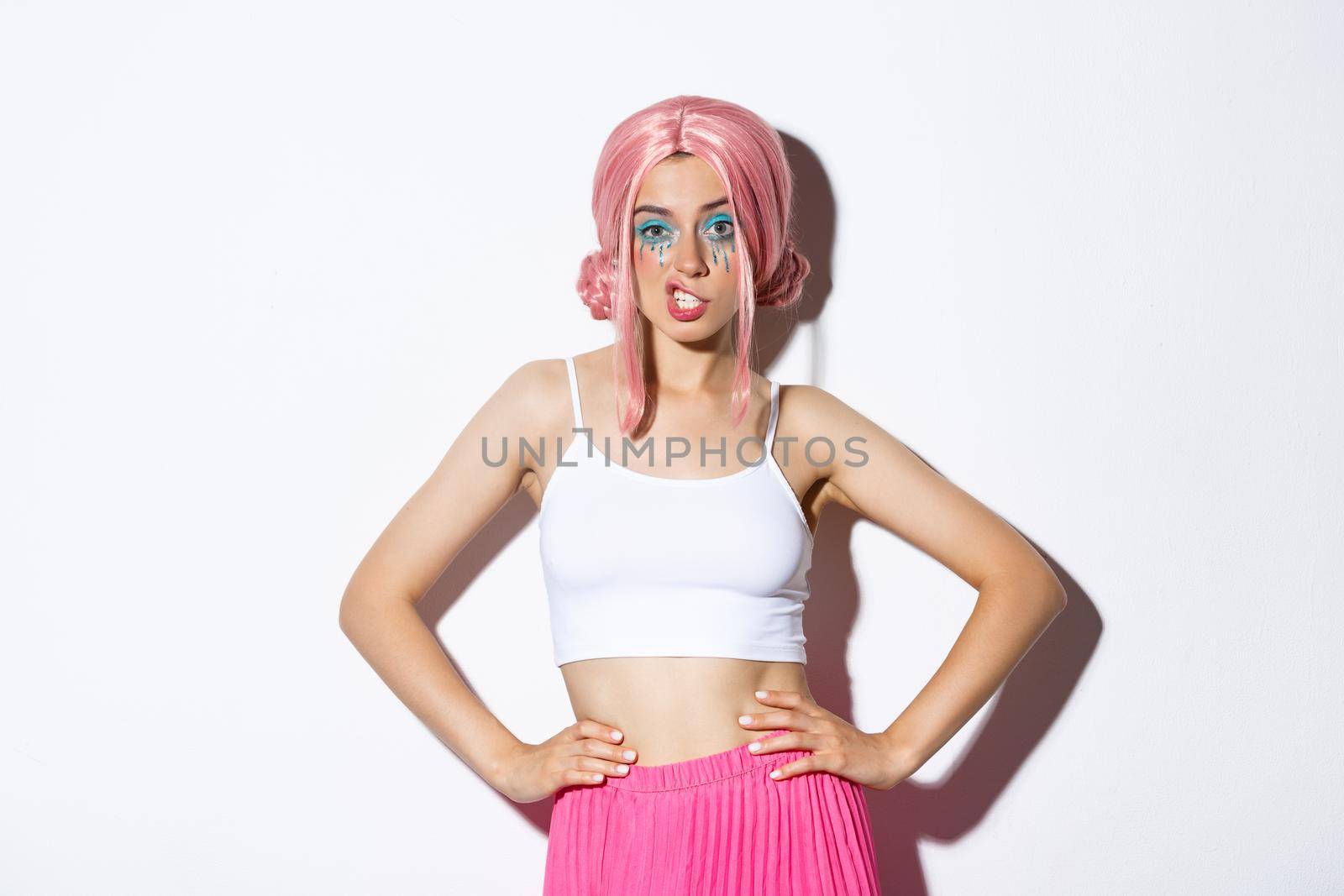 Portrait of sassy attractive woman with pink anime wig, bright makeup, showing teeth and looking confident, celebrating halloween, standing over white background.