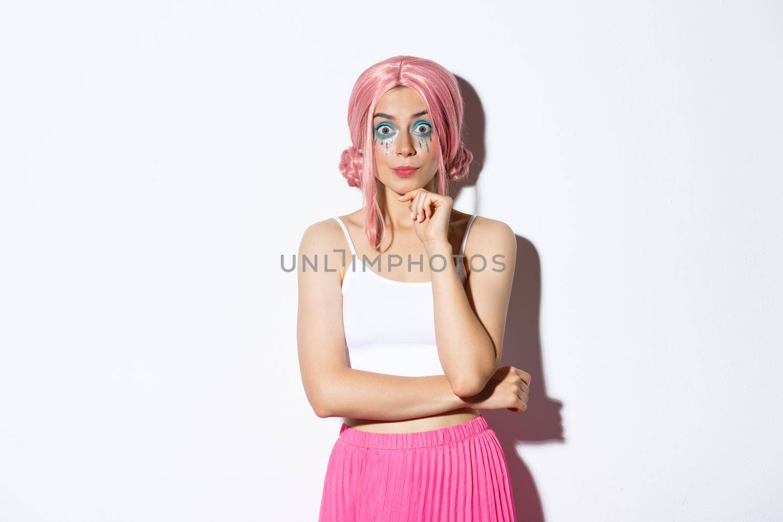 Excited cute party girl with pink wig and halloween makeup, looking with interest at camera, listening to something, standing over white background.