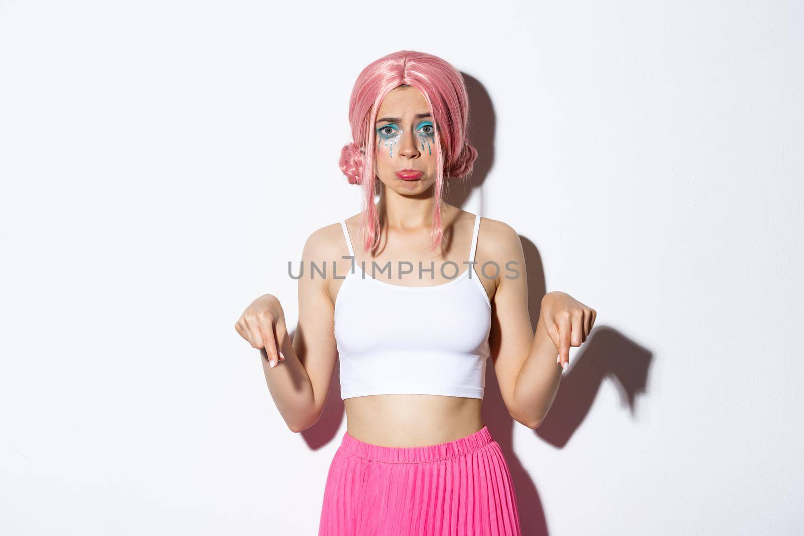 Portrait of disappointed cute sulking girl in pink wig and halloween outfit, looking upset while pointing fingers down, complaining, standing over white background.