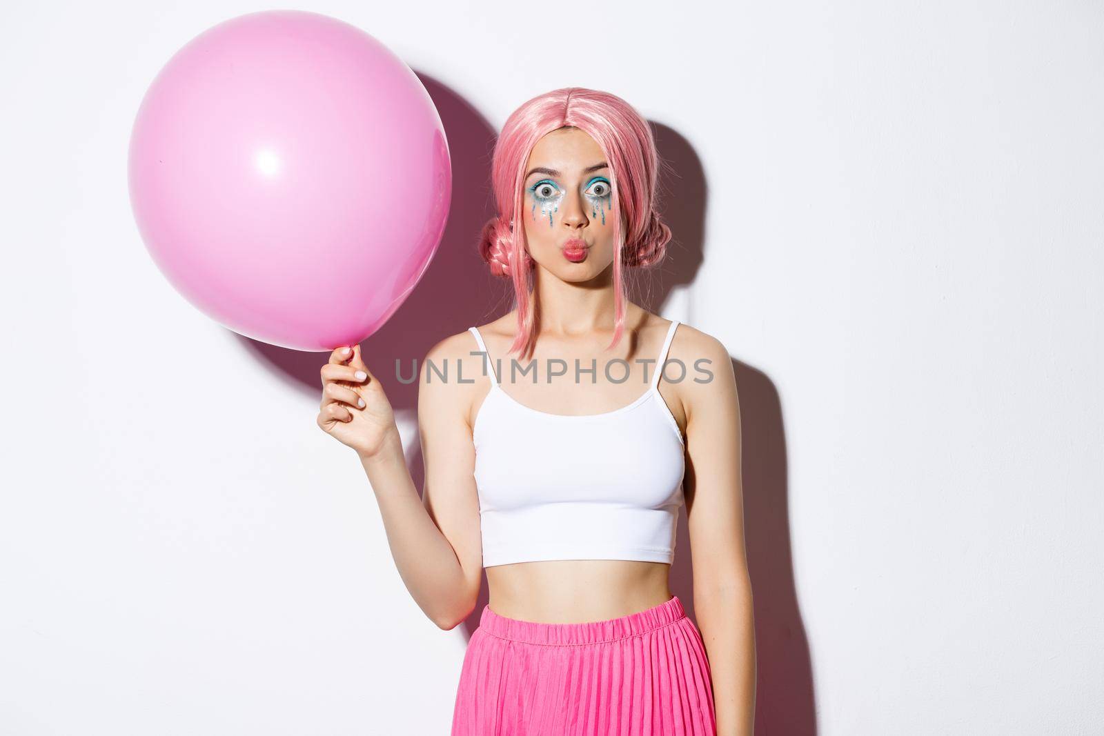 Image of silly party girl with bright makeup, pink wig, holding big cute balloon and pouting, standing over white background.