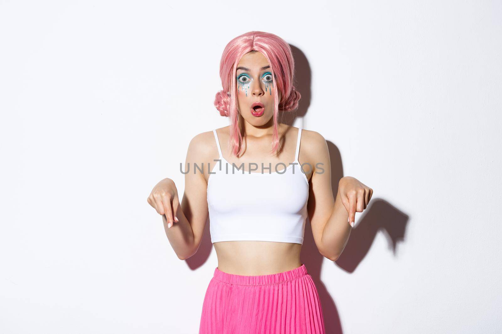 Shocked caucasian girl with pink wig and bright makeup gasping, looking scared and pointing fingers down at halloween banner, standing over white background.