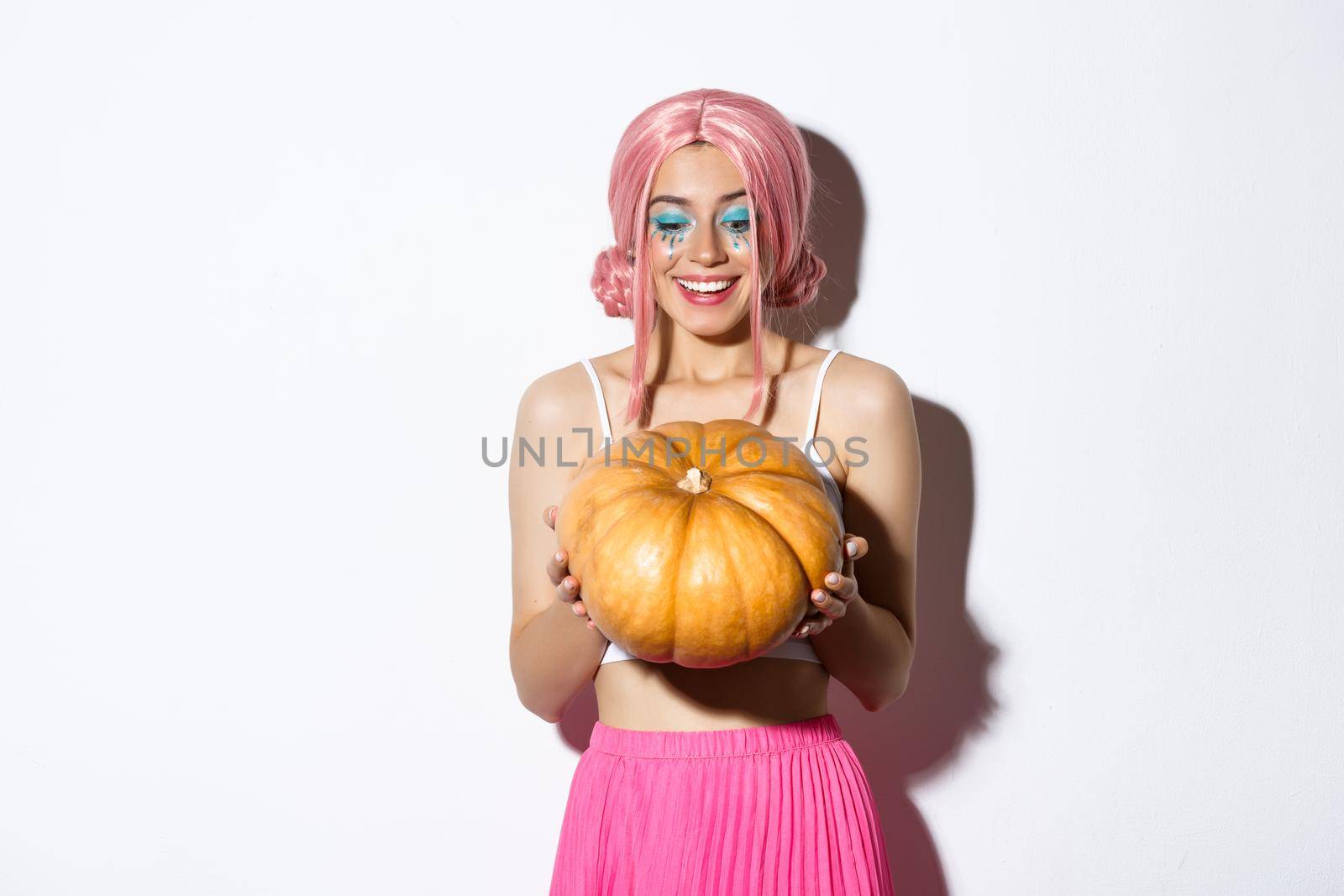 Portrait of cheerful woman with pink wig and bright makeup, looking happy at pumpkin for halloween, standing over white background.