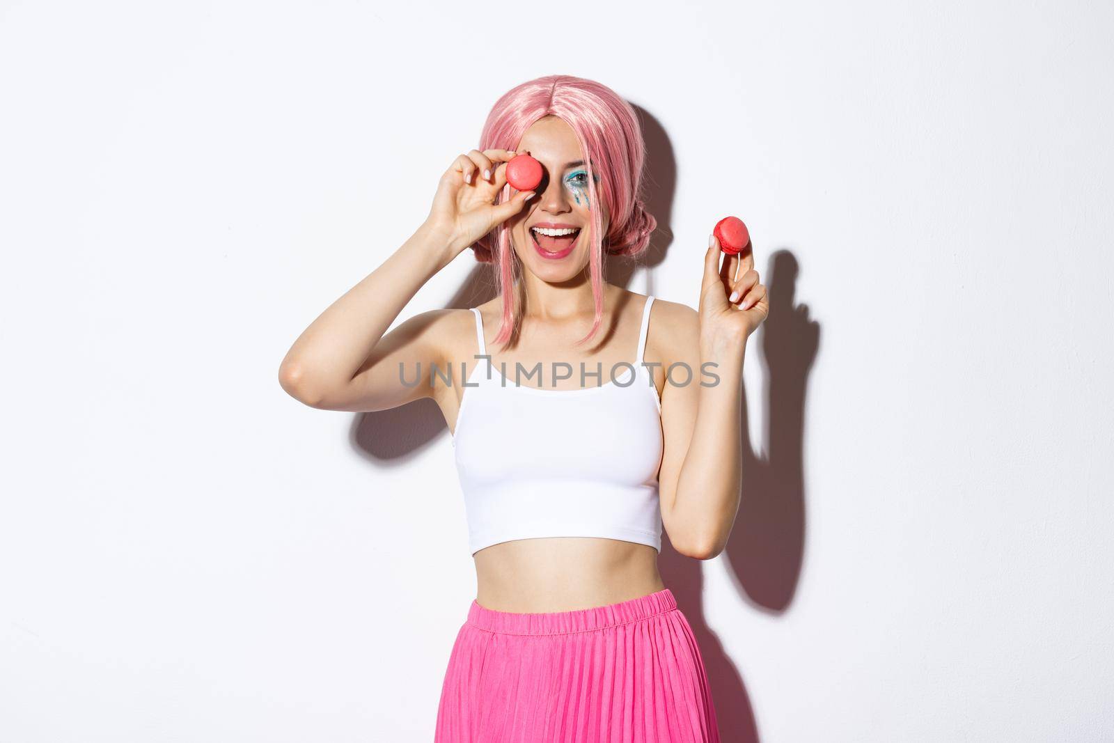 Portrait of cheerful attractive woman in pink wig, celebrating holiday, holding macaroons and smiling, standing over white background.