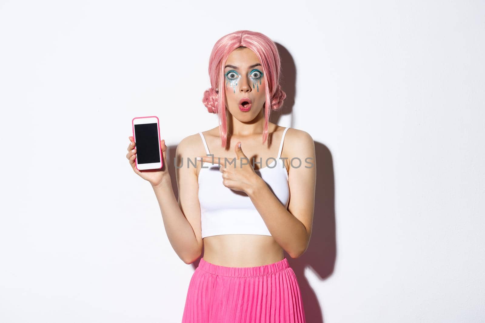 Portrait of surprised beautiful girl in halloween costume, pink wig and bright makeup, pointing finger at smartphone screen, standing over white background.