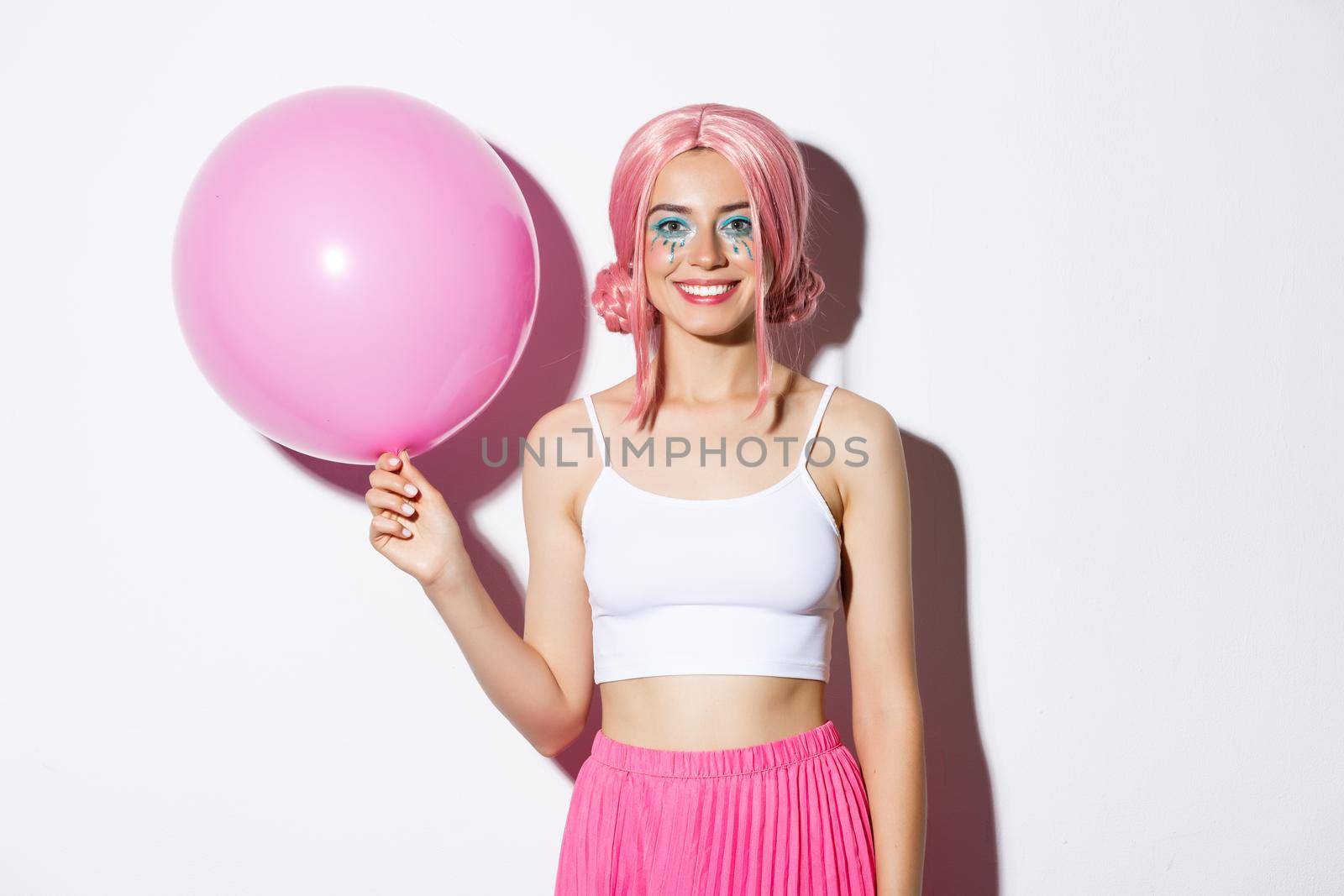 Pretty smiling girl wearing pink wig and holding balloon at holiday celebration or a party, standing happy over white background.