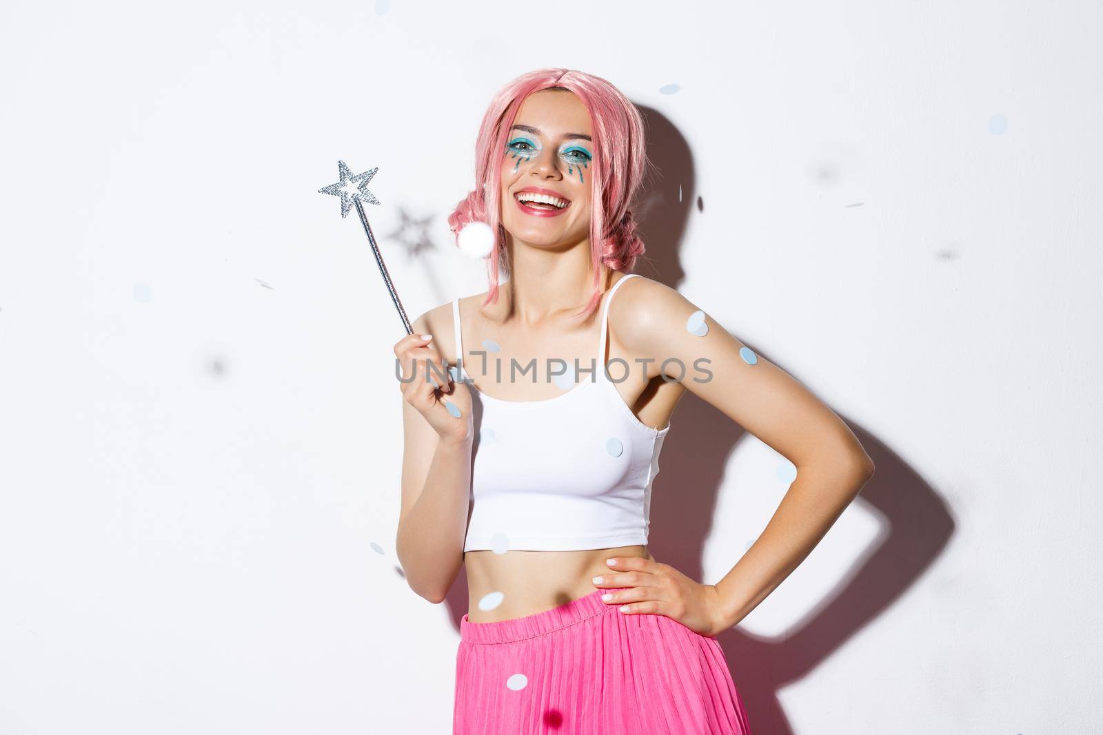 Image of beautiful happy girl in pink wig, celebrating halloween in fairy costume, holding magic wand and smiling while confetti floating in air.