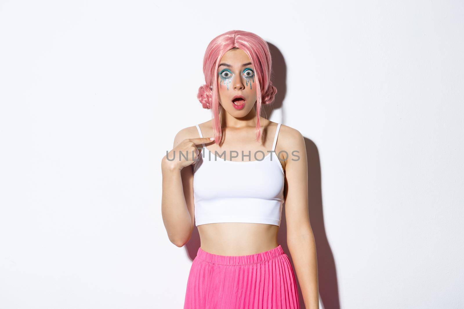 Image of surprised silly girl with pink anime hair and bright makeup, pointing at herself with opened mouth and confused expression, standing over white background.