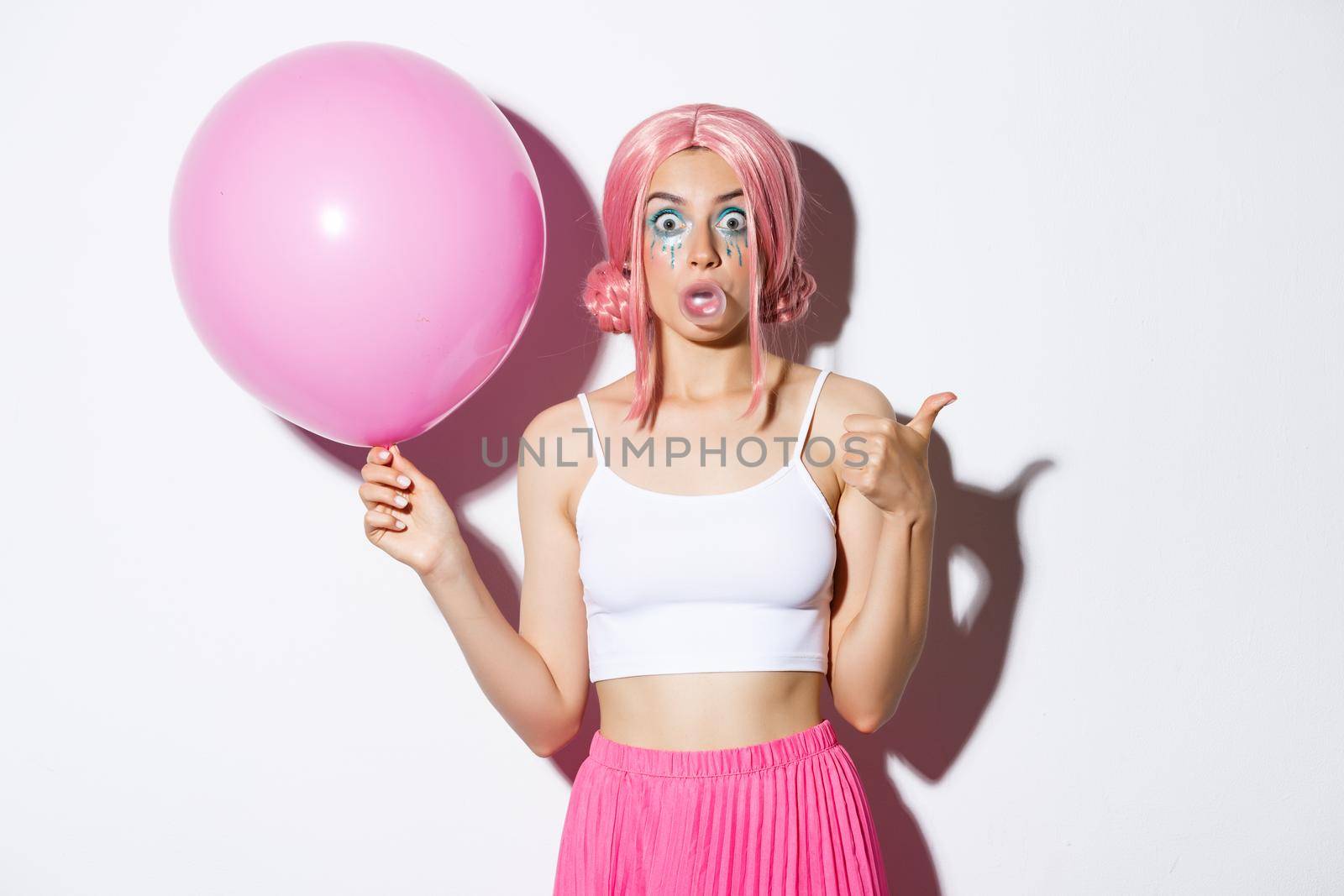 Image of excited attractive girl blowing bubble gum, showing thumbs-up in approval, standing with cute pink balloon while wearing party outfit and wig.