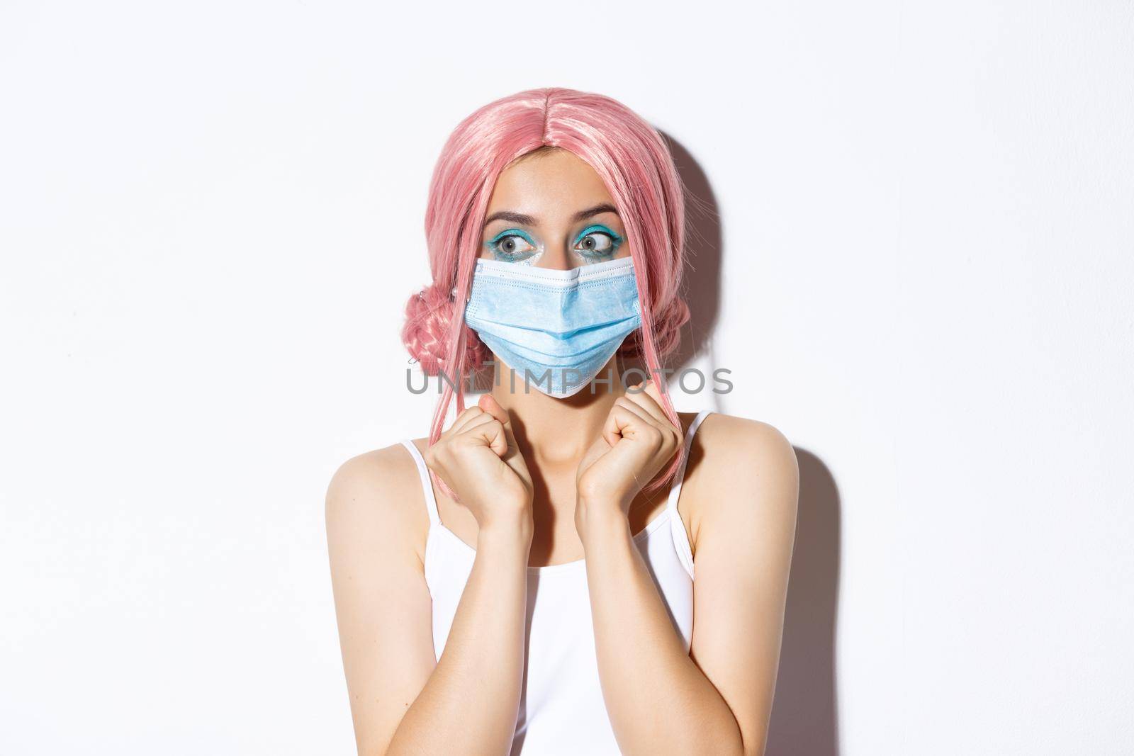 Coronavirus, social distancing and lifestyle concept. Close-up of beautiful excited girl looking left and jumping from temptation, wearing pink party wig and medical mask.