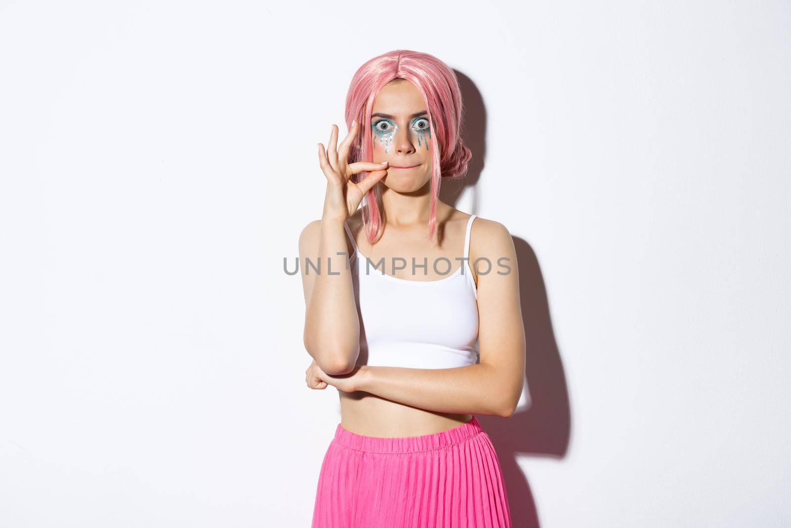 Image of funny party girl in pink wig and halloween makeup, making serious face while making seal on lips, zipping mouth as promise to keep secret, standing over white background.