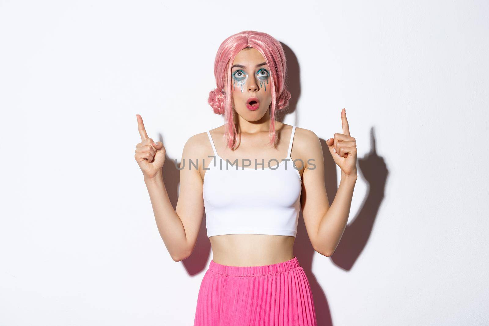 Portrait of surprised cute girl in pink wig, looking and pointing fingers up at promo, standing in outfit for halloween party against white background.