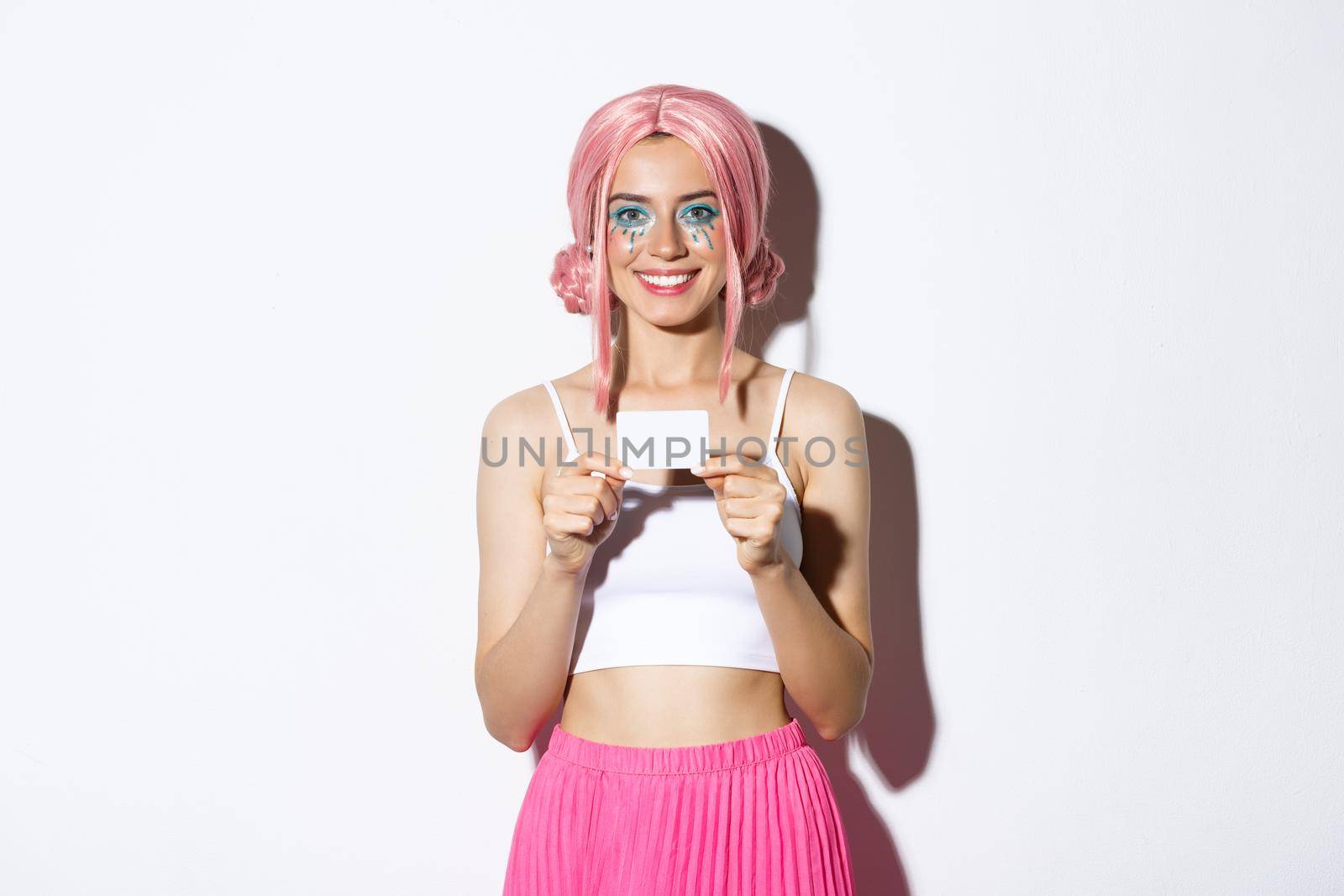 Portrait of beautiful female model wearing pink wig for halloween party, celebrating holiday and showing credit card, standing over white background.
