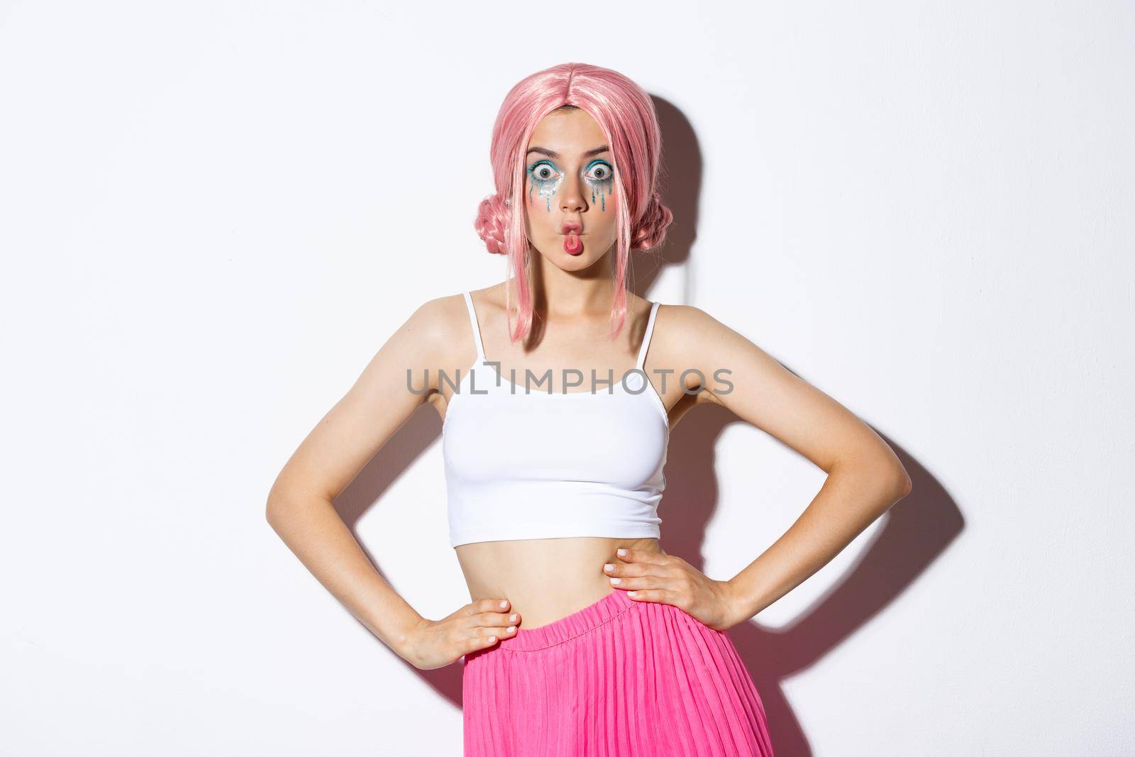 Image of silly party girl with pink wig and bright makeup, celebrating halloween, looking surprised, standing over white background.