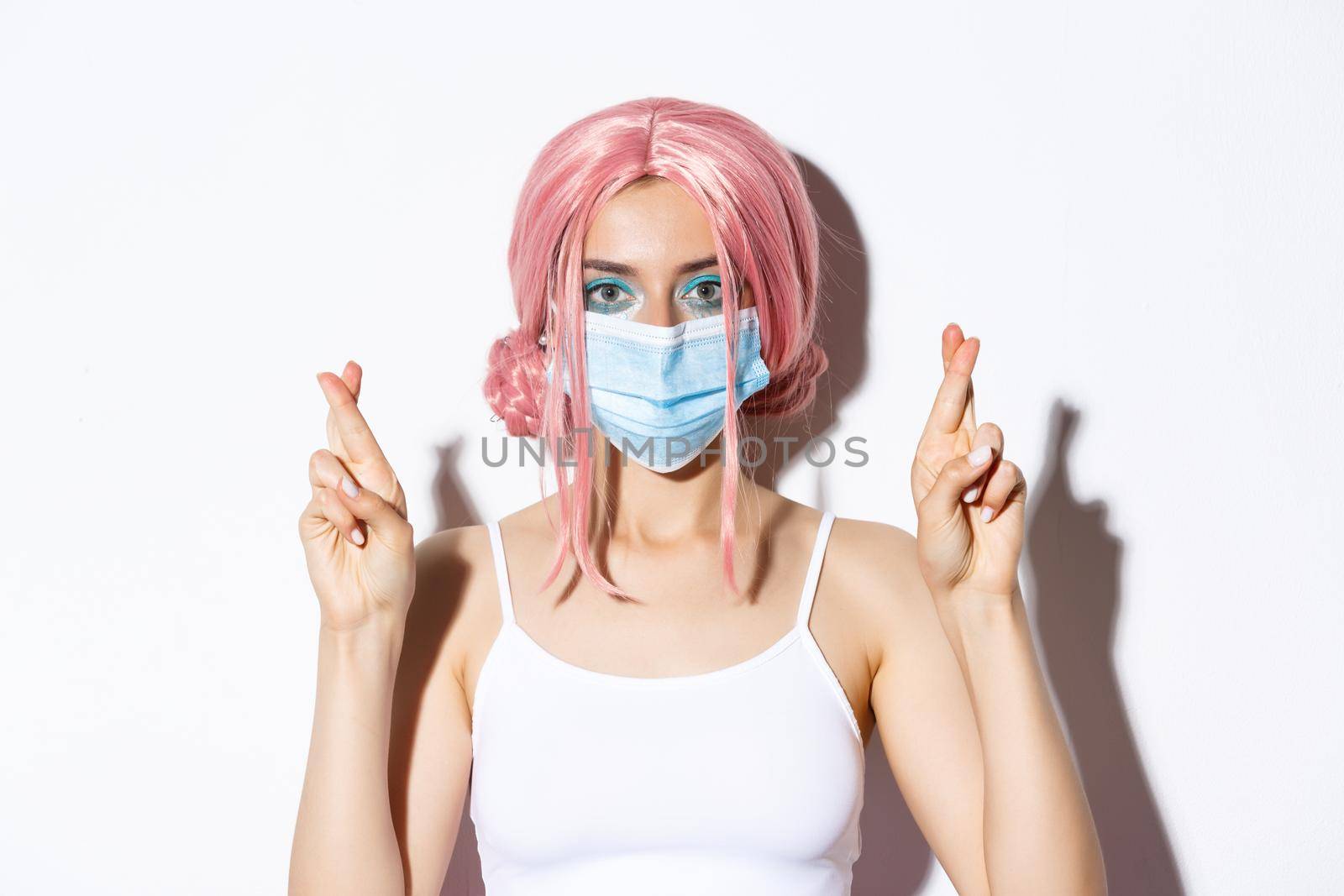 Coronavirus, social distancing and lifestyle concept. Close-up of hopeful girl in pink wig and medical mask cross fingers and making wish.