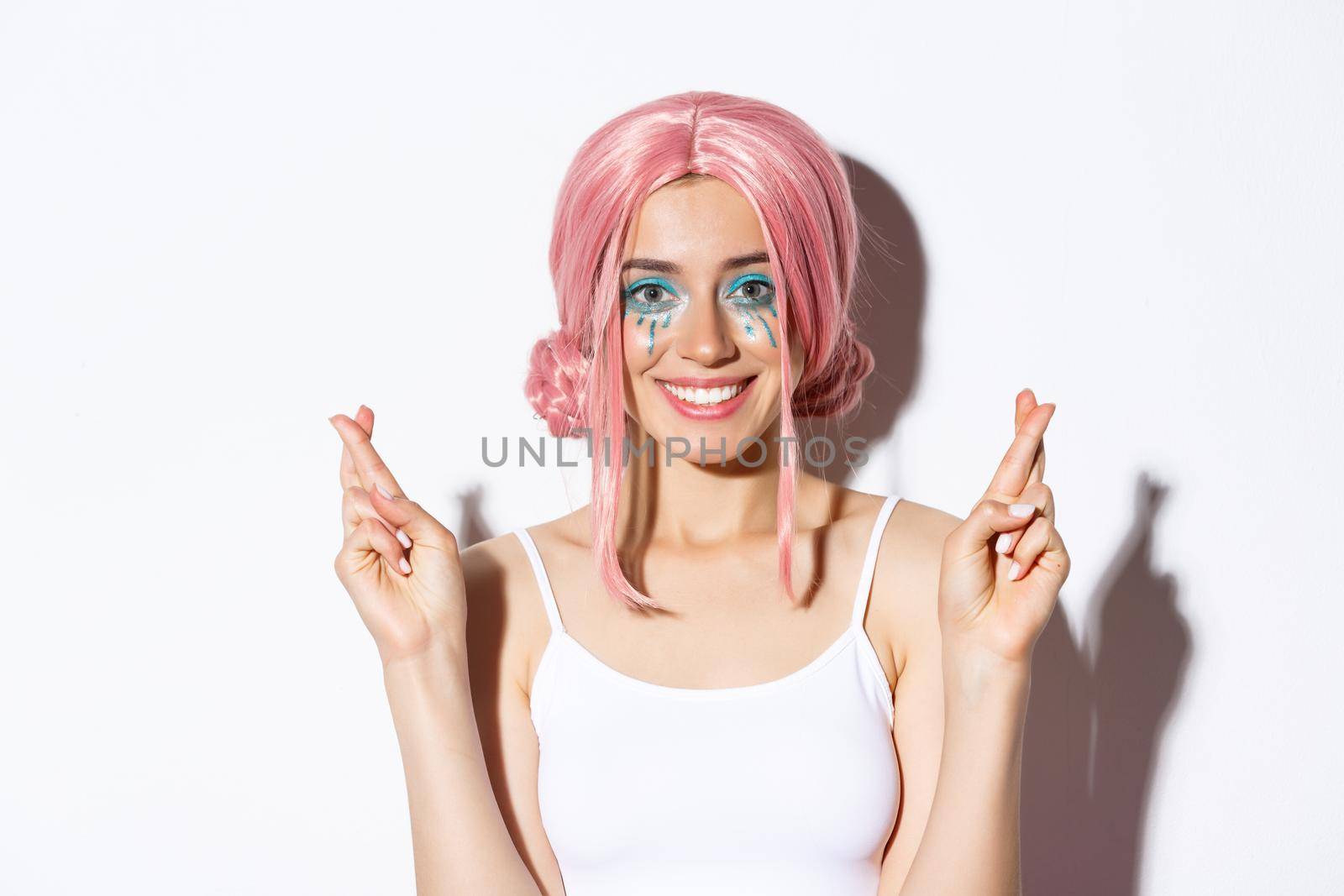 Close-up of lovely smiling woman in halloween costume, pink wig and bright makeup, looking hopeful at camera and making wish with fingers crossed.