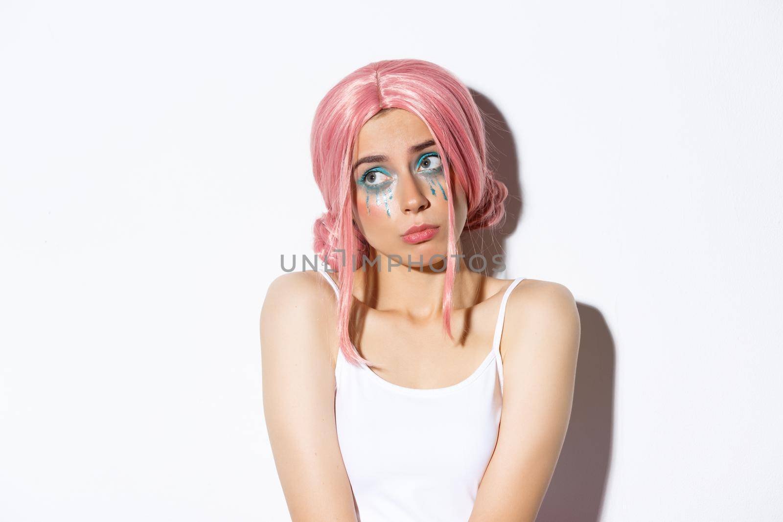 Image of beautiful indecisive girl in halloween costume and pink wig, looking at upper left corner doubtful, standing over white background.