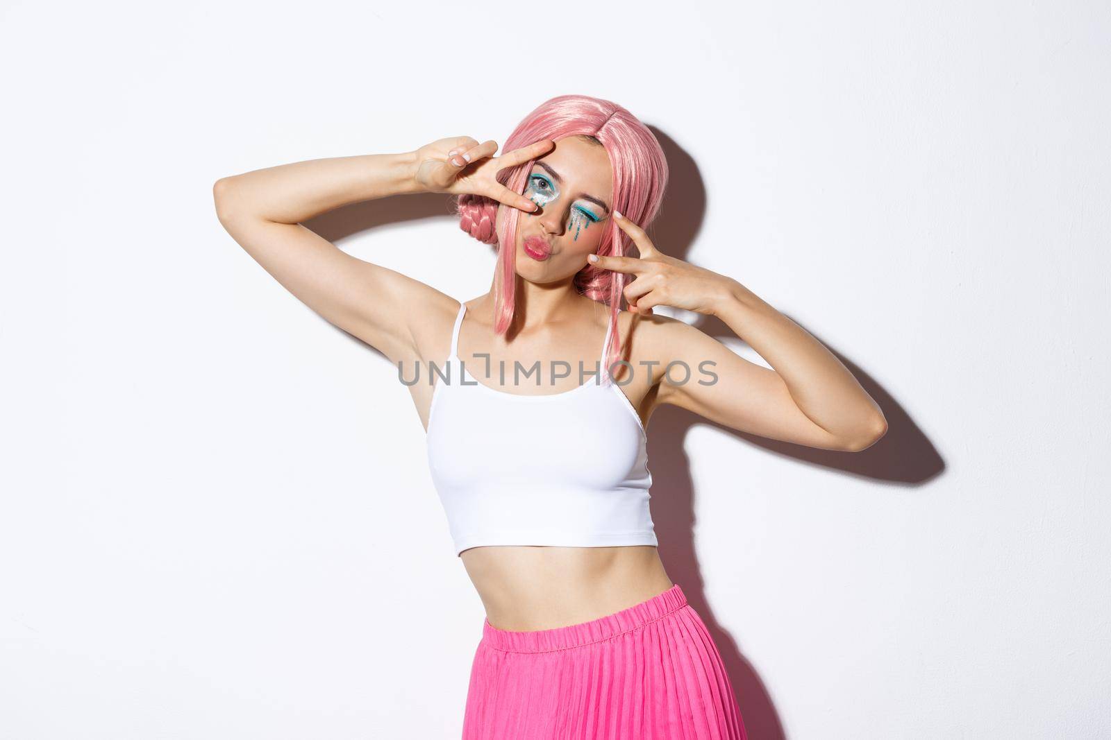 Carefree stylish girl with pink wig and bright makeup, having fun on party, celebrating and dancing, showing peace signs, standing over white background.