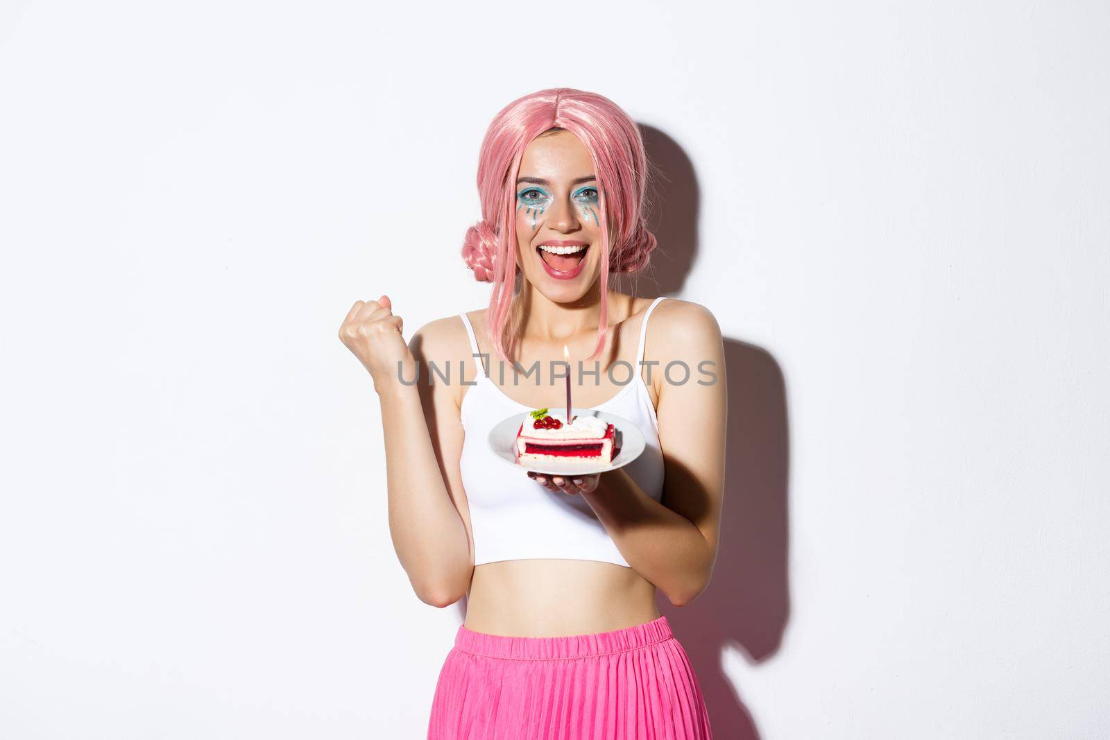 Portrait of cheerful smiling girl celebrating her birthday, wearing pink wig, holding b-day cake and shouting of joy, standing over white background.