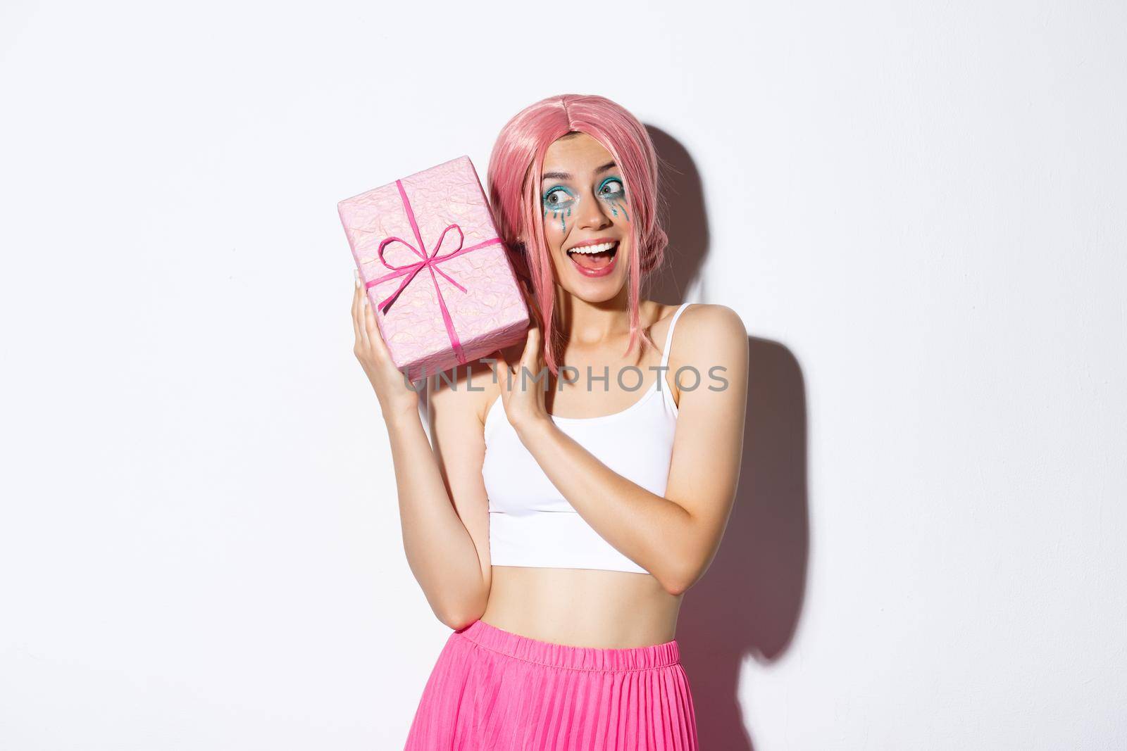 Image of beautiful girl in pink wig shaking box with birthday gift, wonder what inside wrapped box, standing over white background.