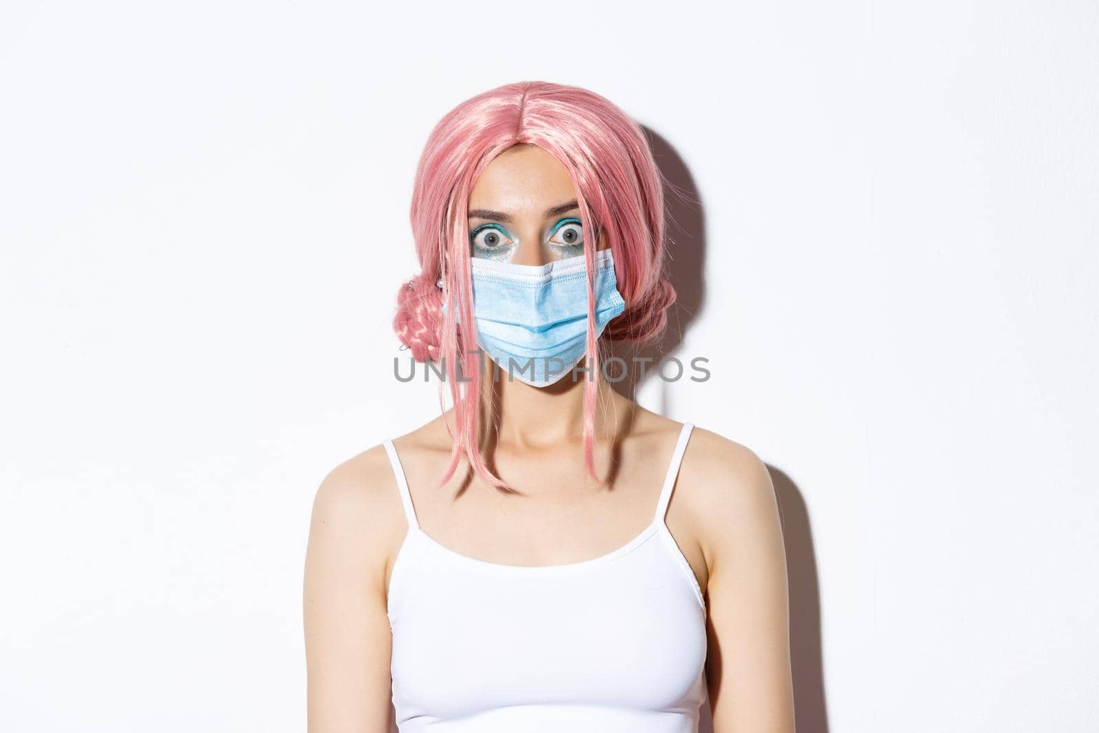 Close-up of surprised girl in pink wig and medical mask looking at camera amazed, protect herself against coronavirus disease, standing over white background.