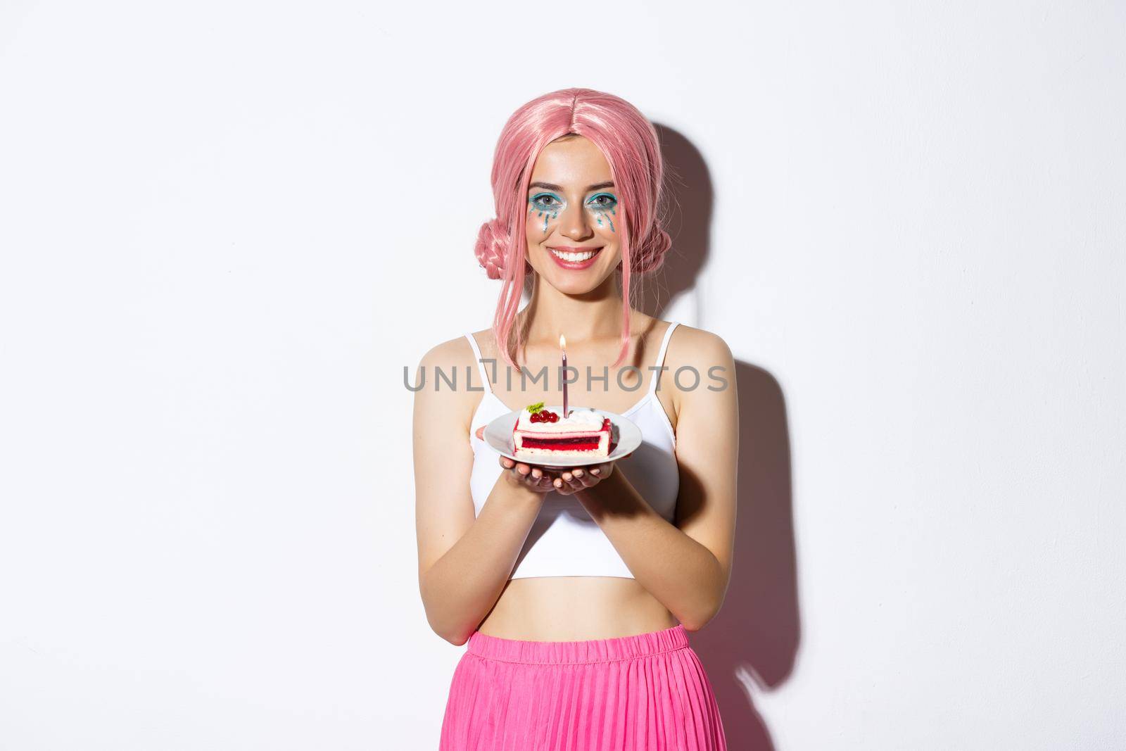 Image of beautiful birthday girl in pink wig, holding b-day cake with lit candle, smiling and making wish, celebrating holiday over white background.