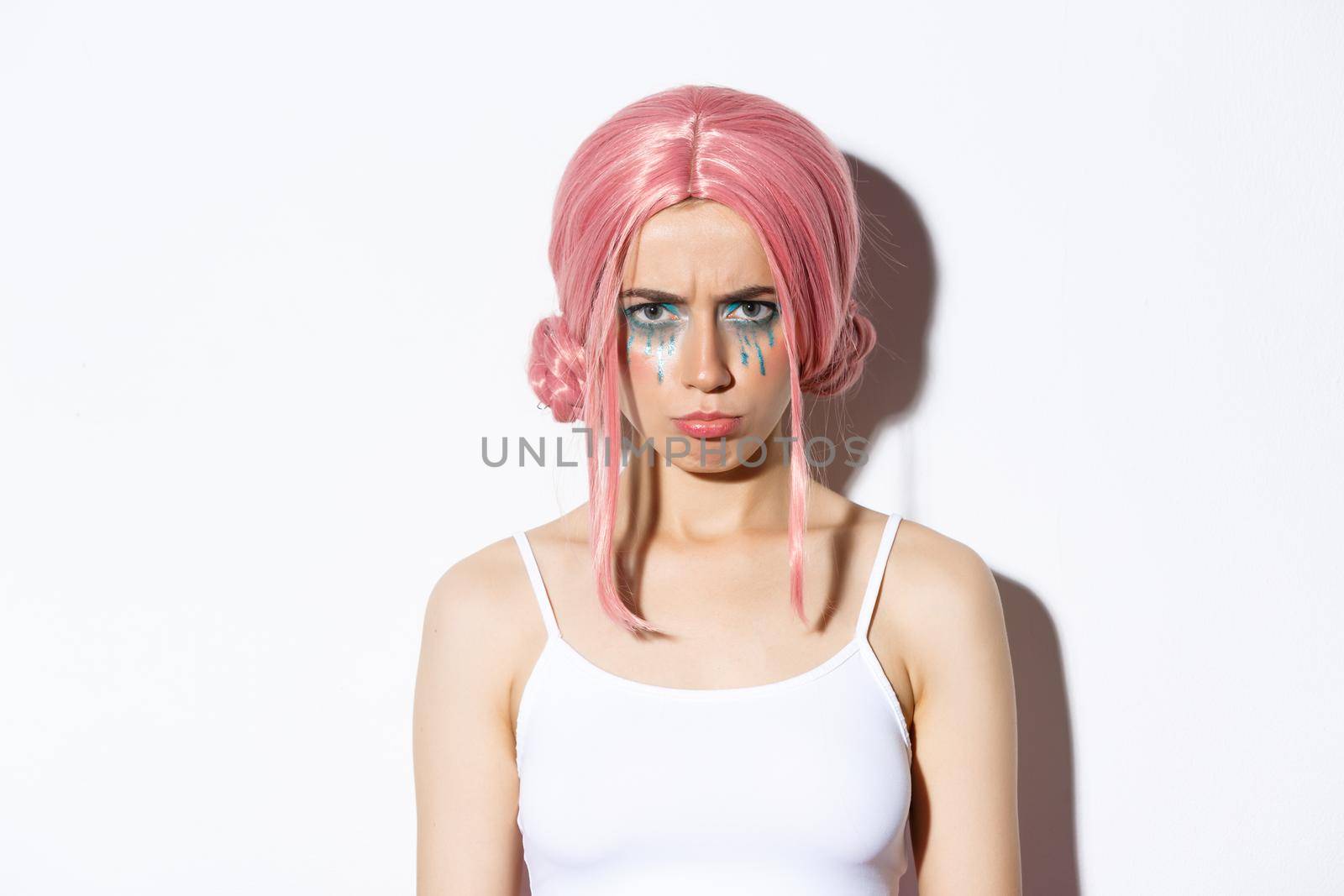 Close-up of angry cute girl with pink wig and glamour makeup, sulking mad at someone, feeling offended, standing over white background.