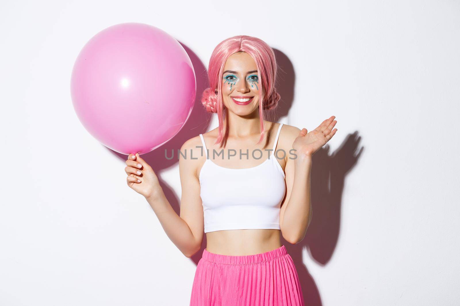 Image of cheerful young girl with pink wig and bright makeup, holding balloon and waving hand to say hi, greeting someone at party, celebrating holiday, white background.