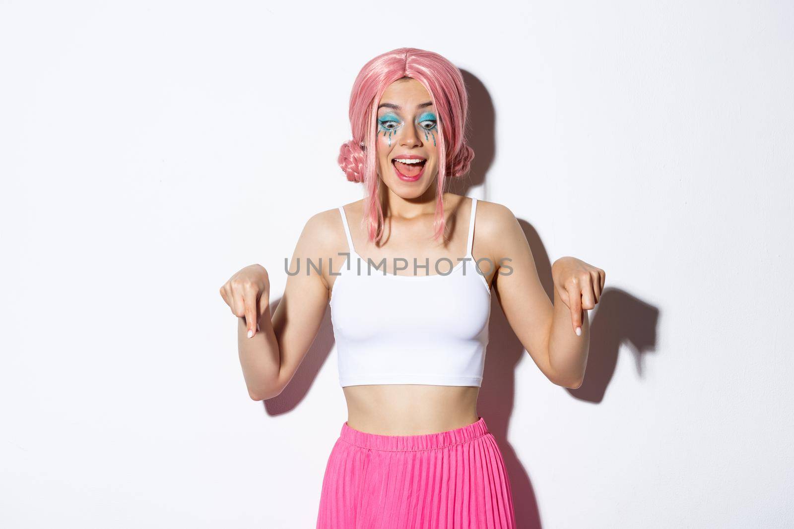Portrait of excited party girl in pink wig and bright makeup, react to something amazing, pointing fingers down and gasping fascinated, standing over white background.
