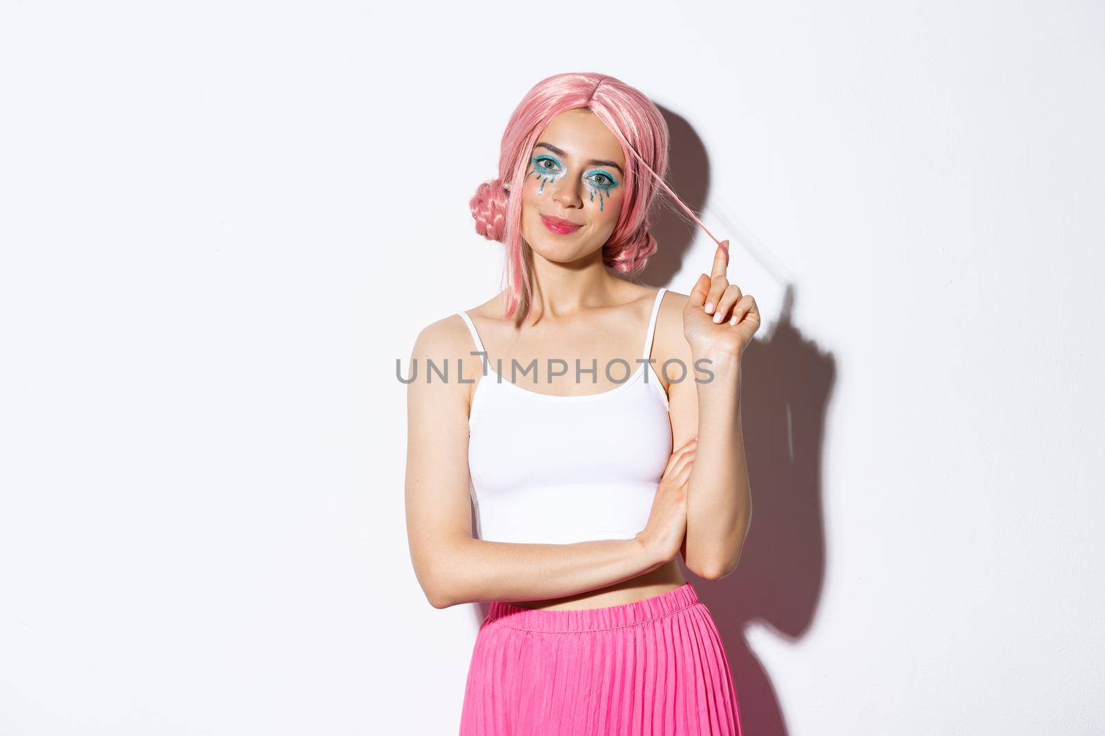 Image of beautiful female model in halloween outfit, wearing pink wig and colorful clothes, smiling at camera, standing over white background.