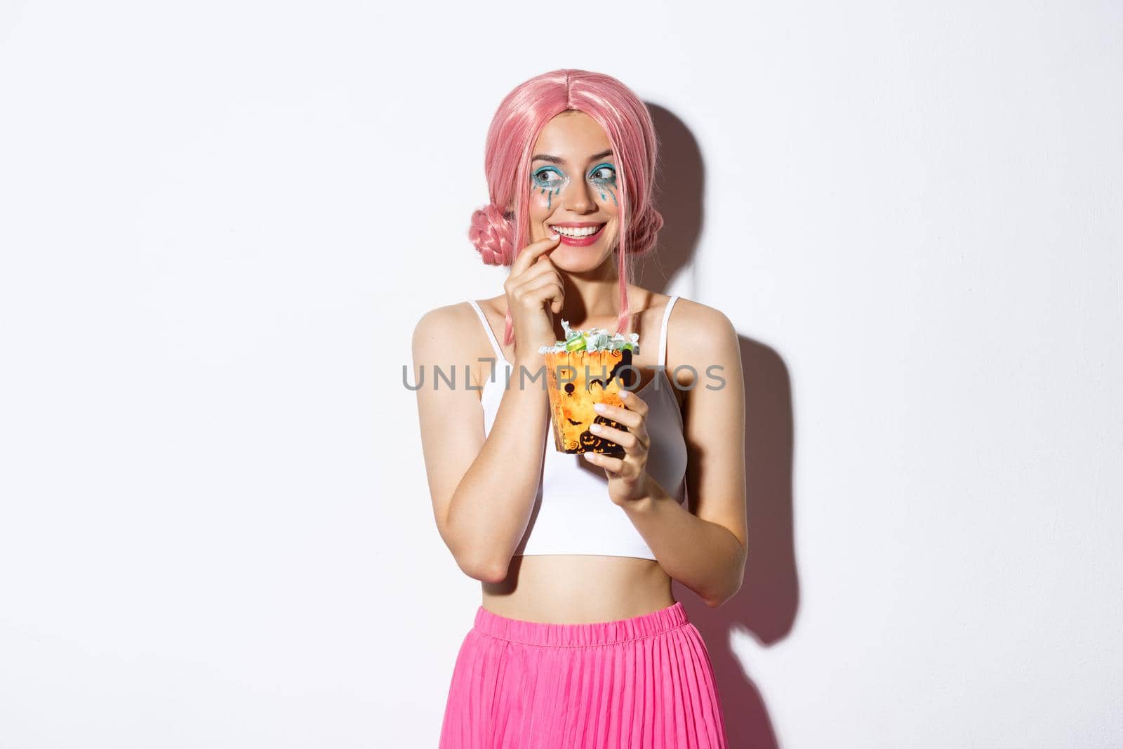 Portrait of thoughtful smiling girl in pink wig, thinking about trick or treat while holding candies, celebrating halloween, standing over white background.