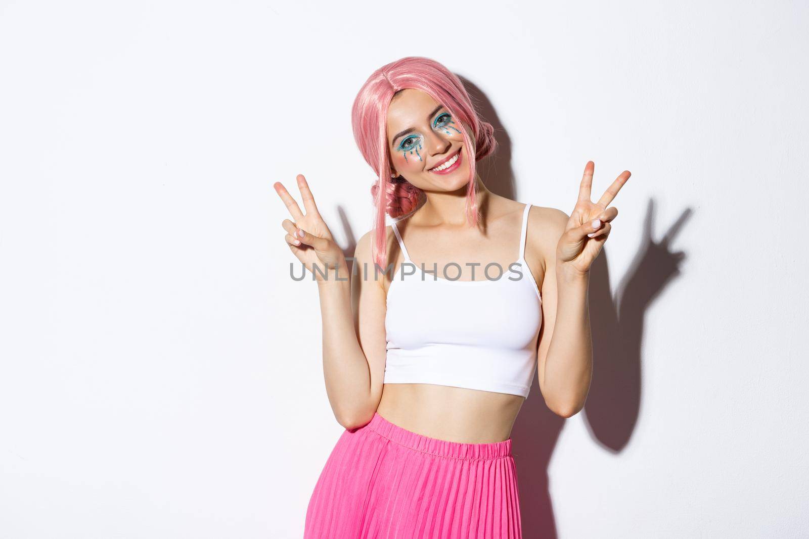 Beautiful girl with lovely smile, showing peace signs, wearing pink wig and halloween makeup, standing over white background.