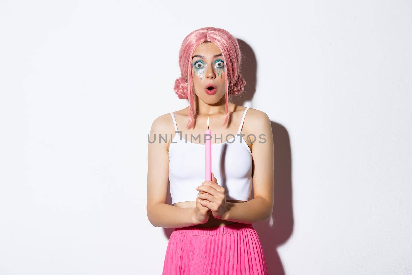 Portrait of shocked girl casting magic spells on halloween, holding candle and looking amazed, standing in costume with pink wig and bright makeup.
