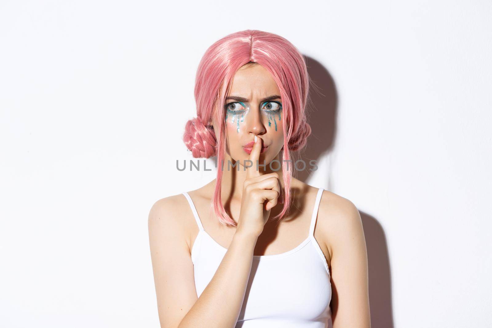 Close-up of worried attractive girl in pink anime hair, showing shush sign, press index finger to lips and looking concerned. Woman share a secret, standing over white background.