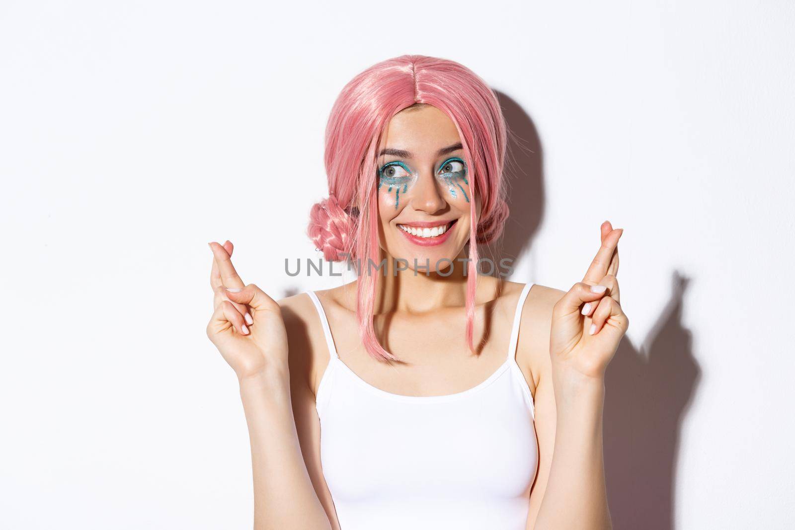 Close-up of lovely hopeful girl in pink wig, cross fingers and making wish, smiling and looking left, anticipating for something, standing over white background.