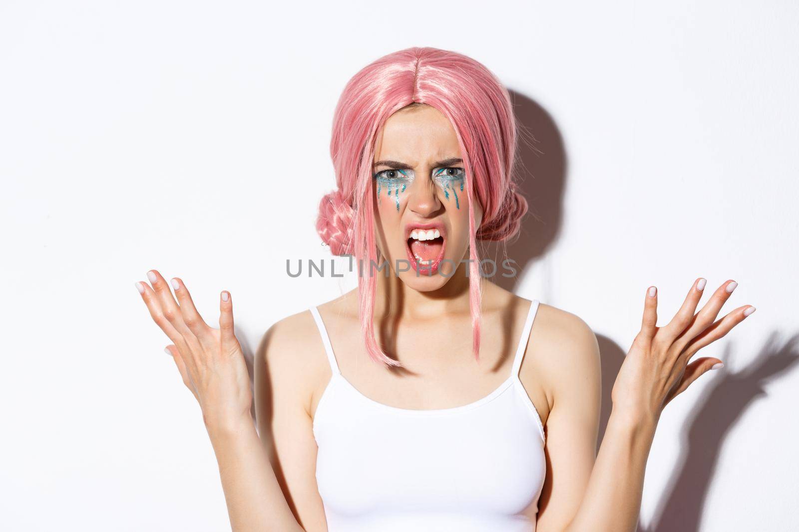 Close-up of angry girl in pink wig, yelling at someone and shaking hands mad, standing over white background.