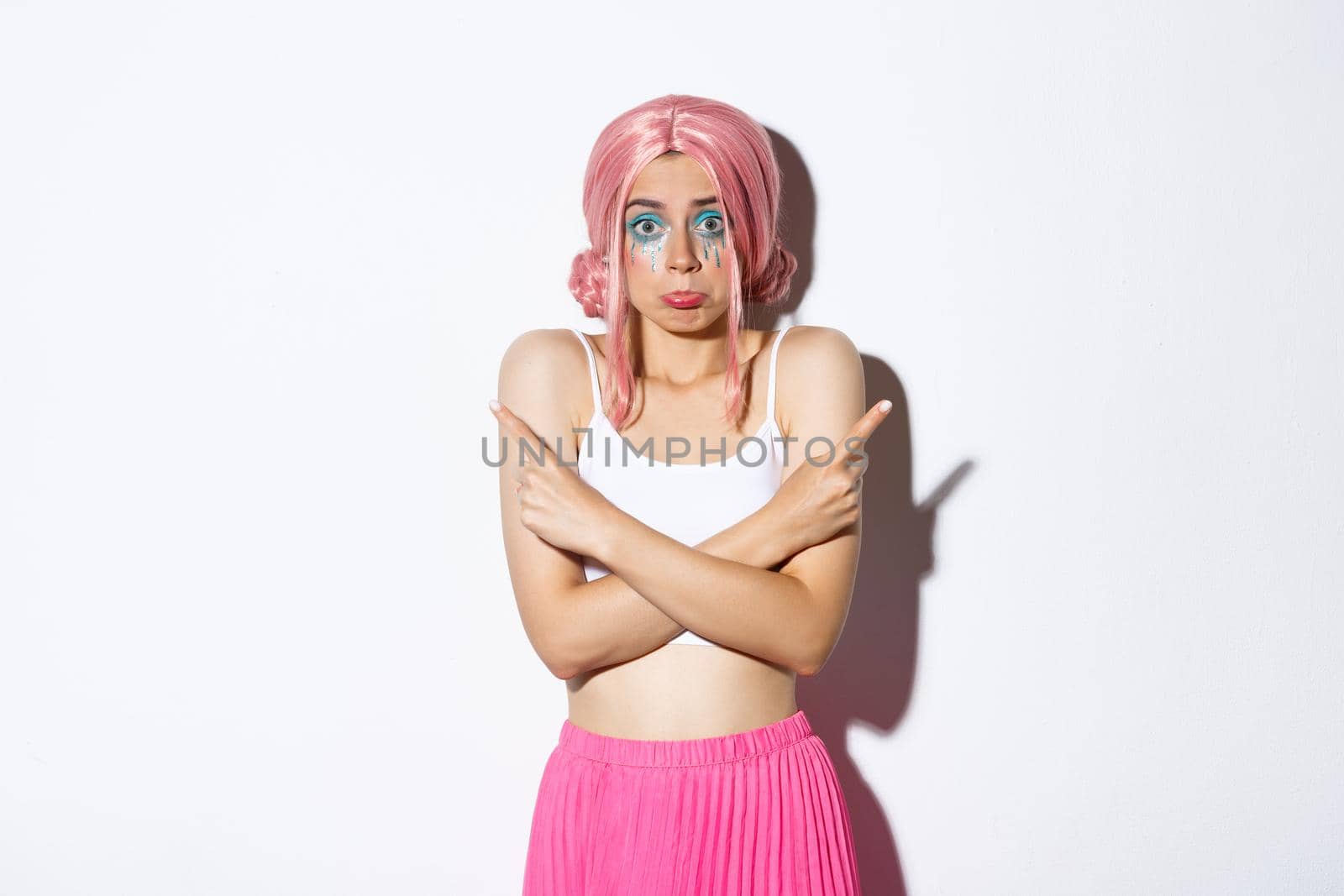 Indecisive silly female model in pink wig, with colorful makeup for halloween party, pointing sideways and shrugging, asking for help with choice, standing clueless over white background.
