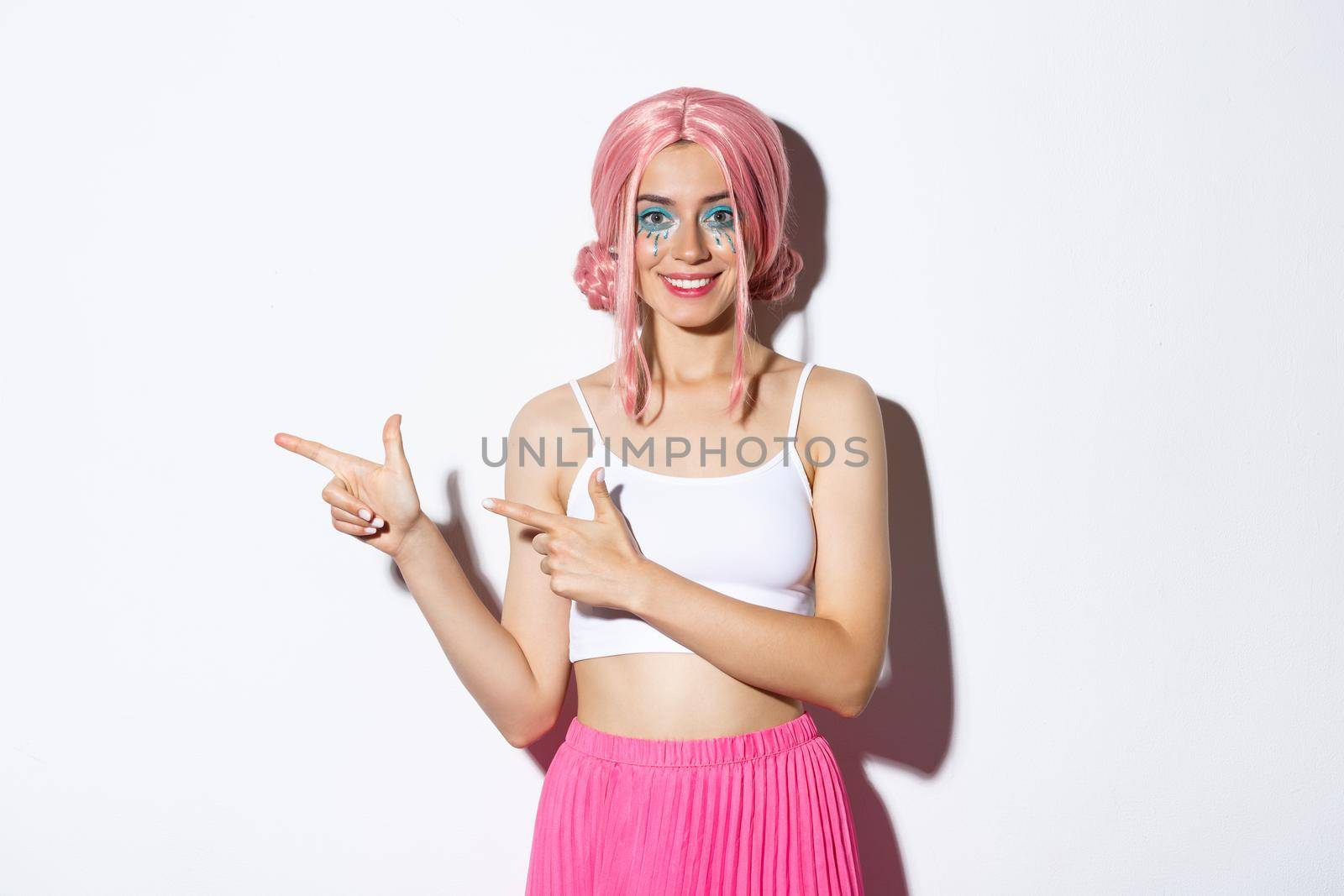Image of trendy smiling girl dressed for halloween party, pointing fingers left, showing advertisement, standing over white background.