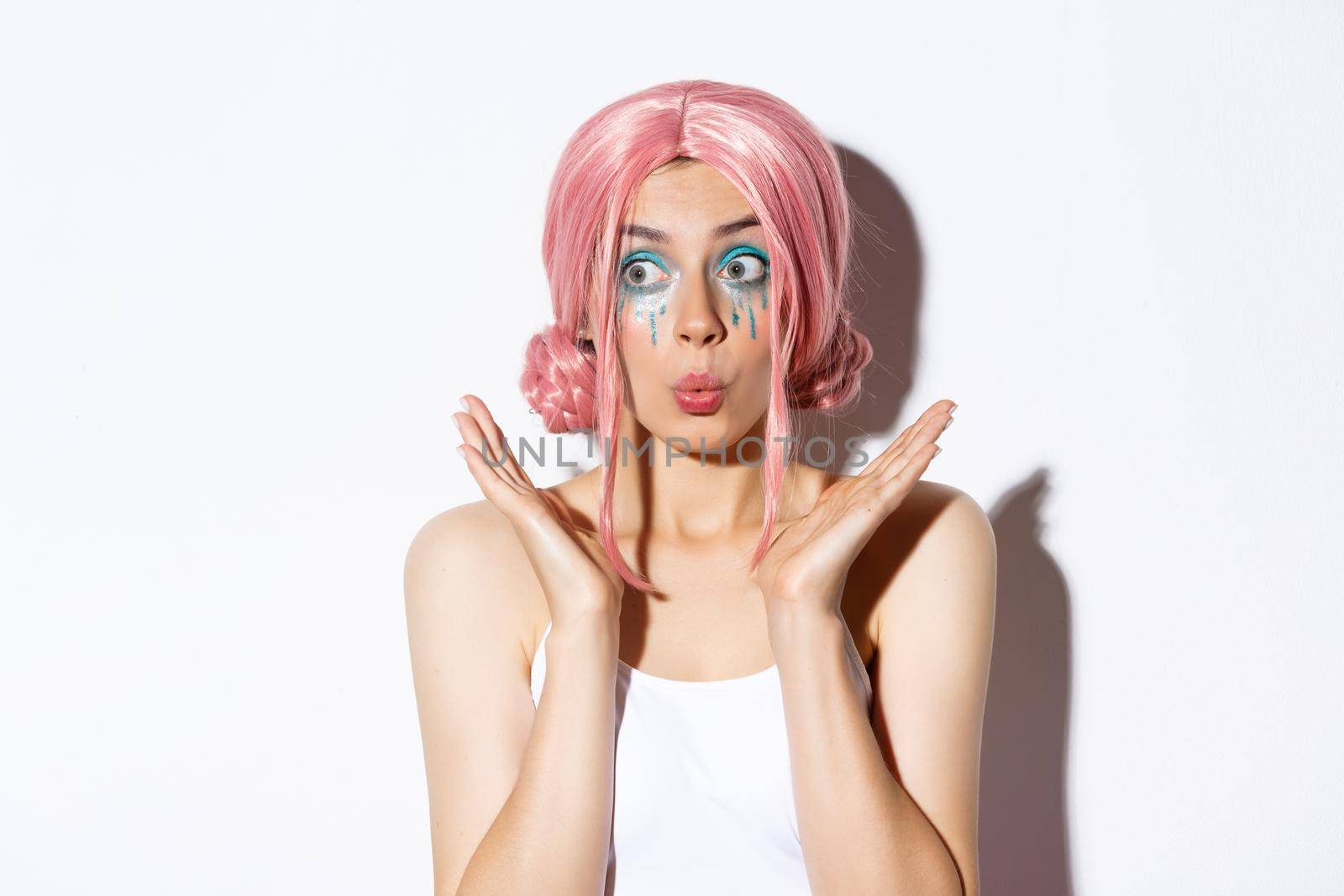 Close-up of excited party girl in pink wig and bright makeup looking impressed, smiling and gazing surprised, standing over white background.