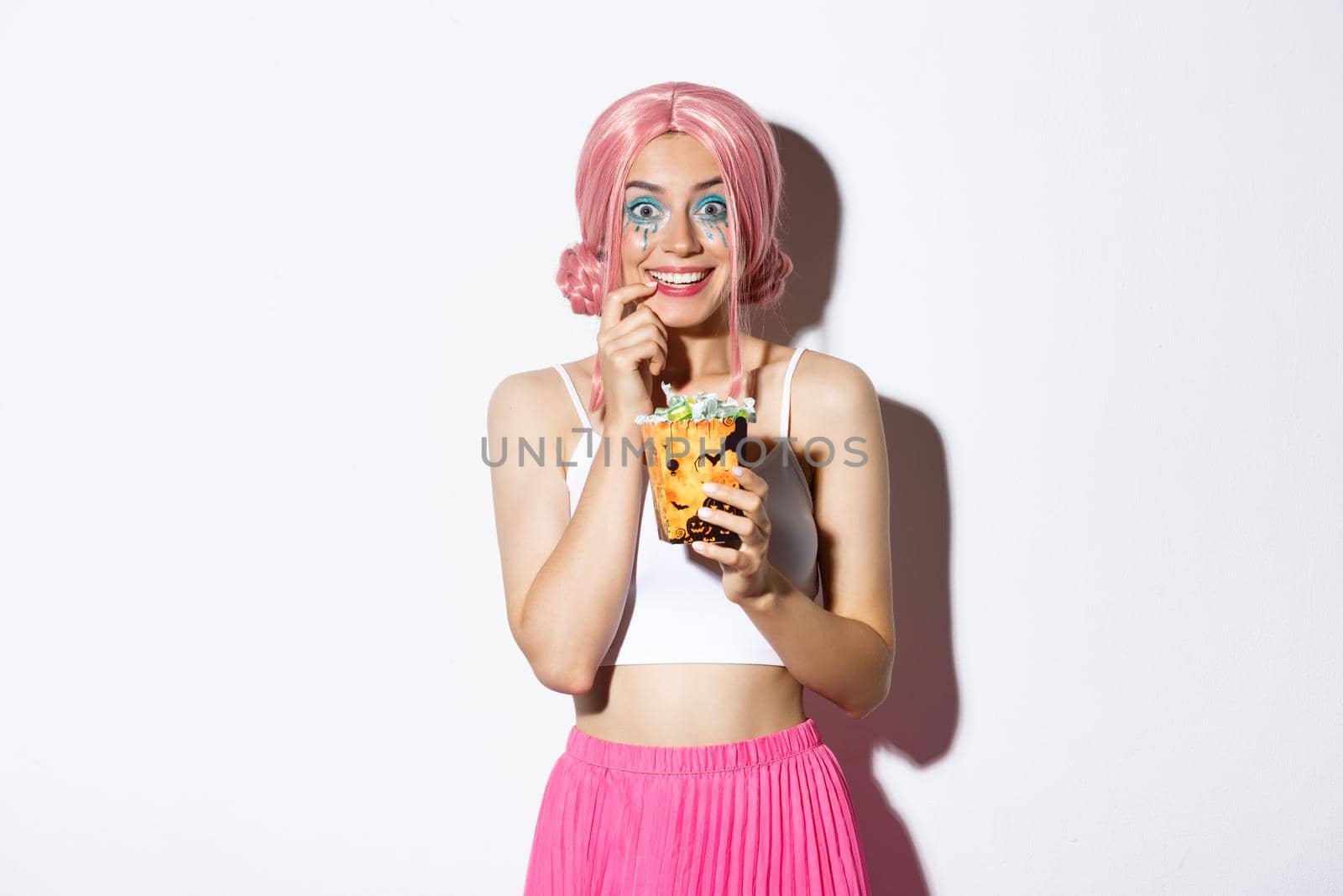 Image of happy attractive girl with pink wig and bright makeup going trick or treat, celebrating halloween, showing candies and smiling, standing over white background.