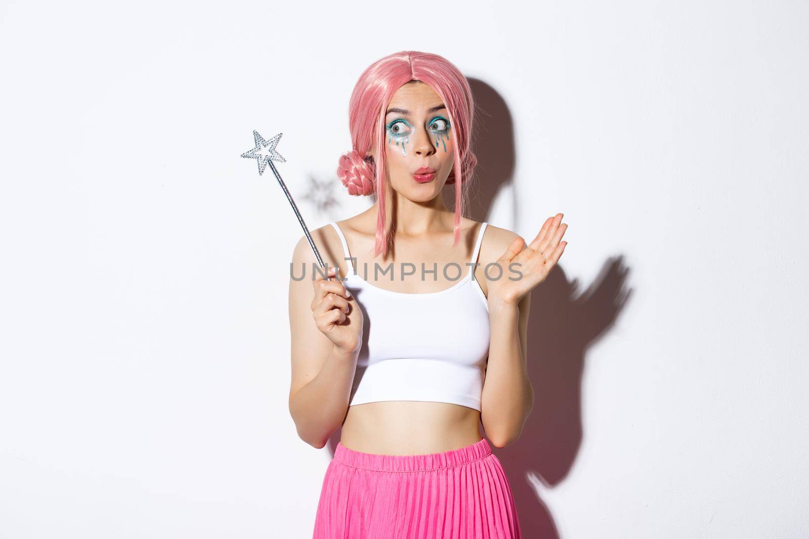 Image of amused cute girl with pink wig and bright makeup, dressed up as fairy for halloween party, holding magic wand and looking excited, standing over white background.