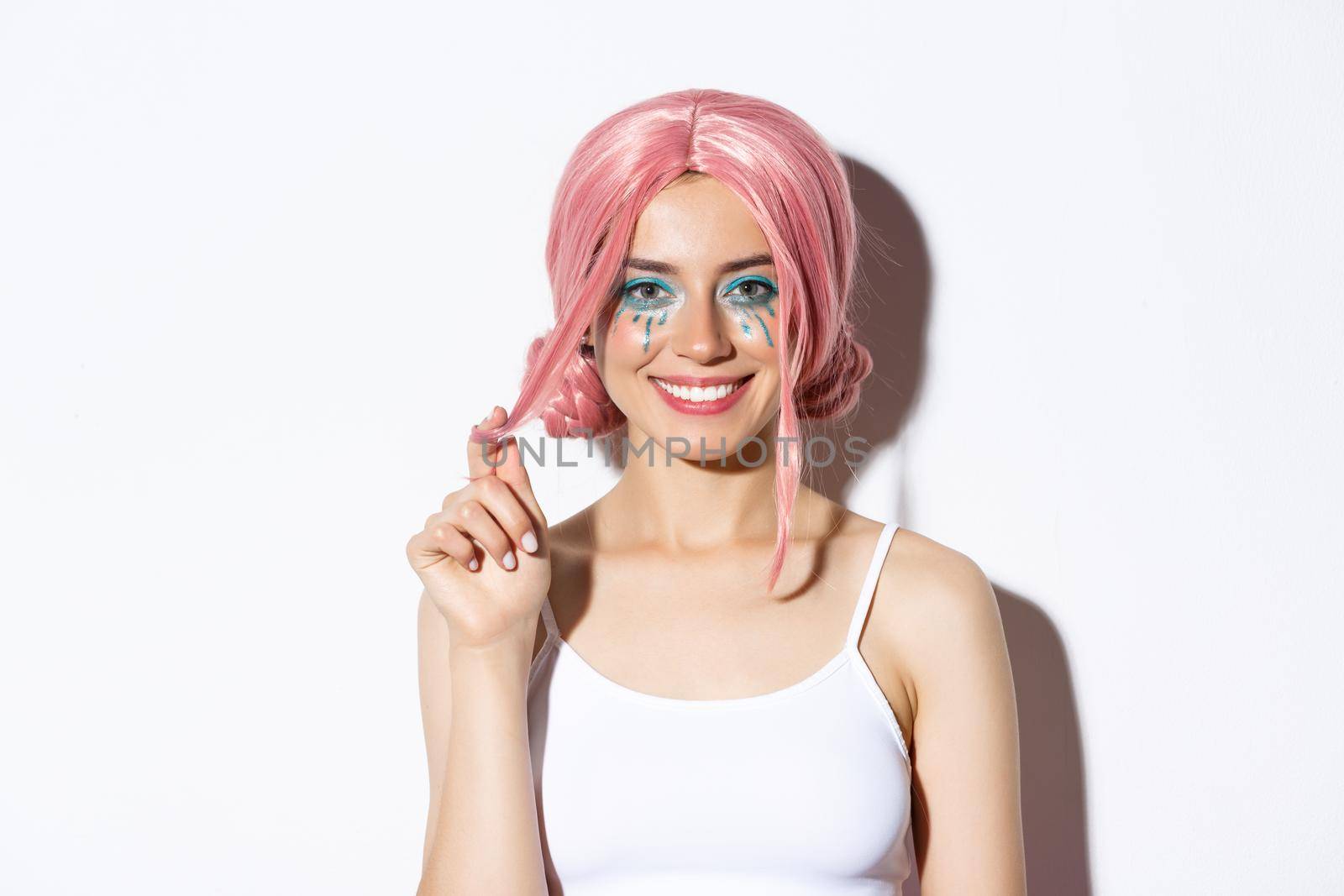 Close-up of beautiful smiling woman in pink wig, celebrating holiday, looking happy at camera, standing over white background.