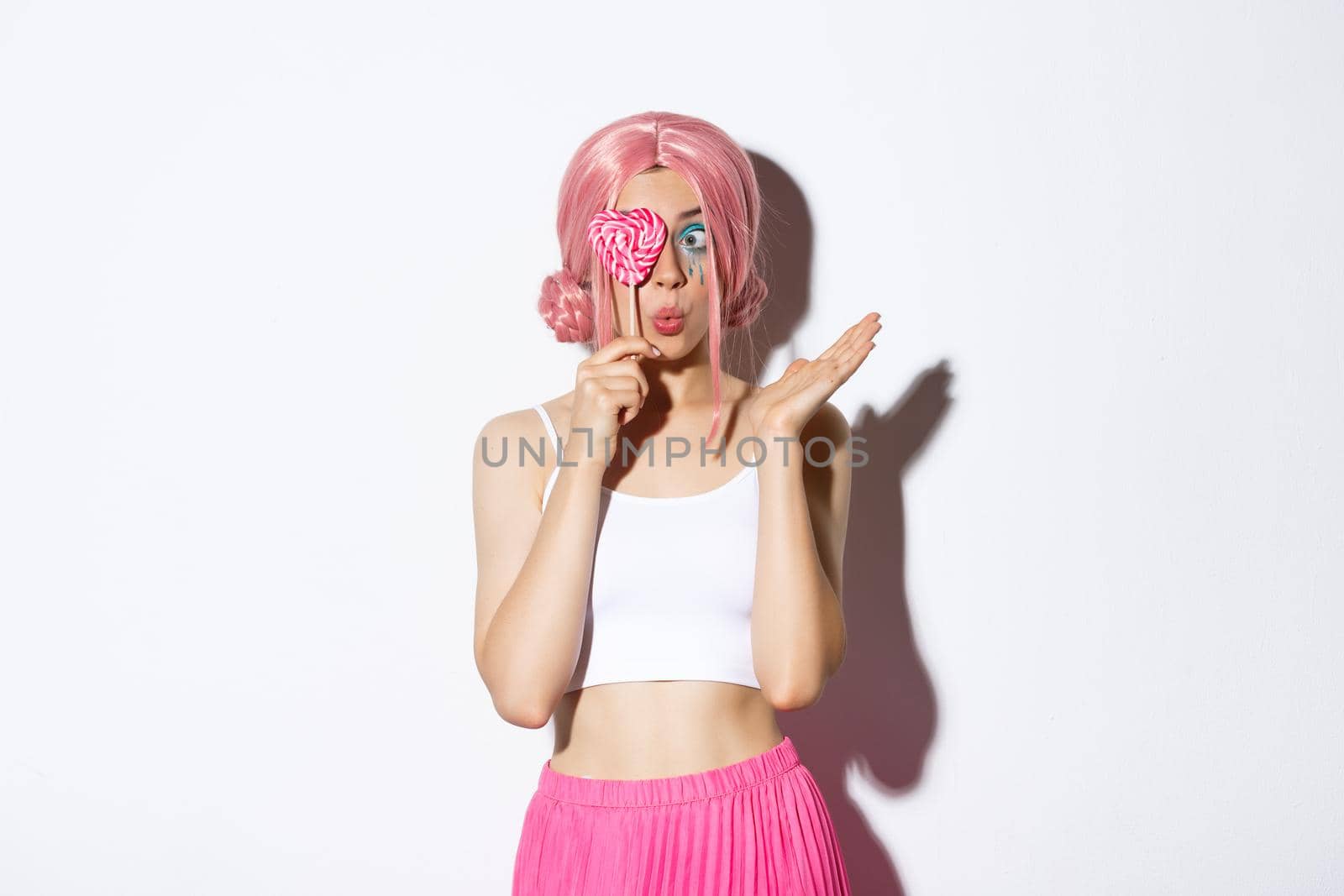 Portrait of attractive girl in pink wig holding cute candy over eye and pouting, wearing fairy costume on halloween, standing over white background.
