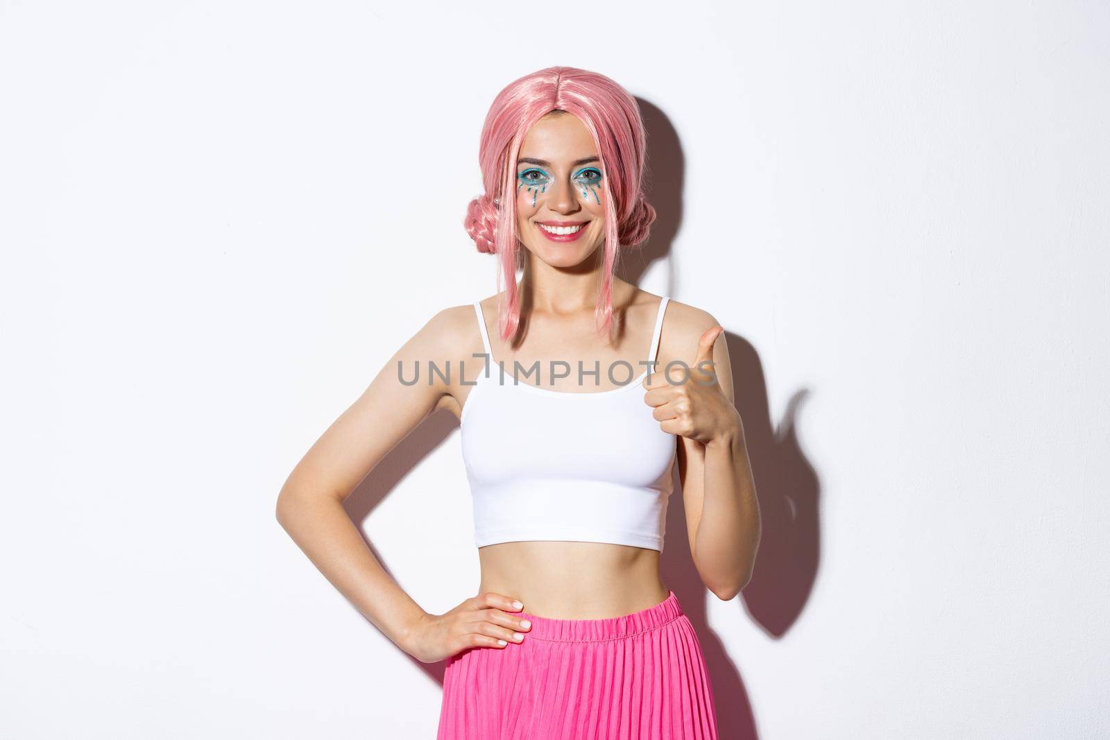 Portrait of confident smiling party girl celebrating halloween, showing thumbs-up in approval, satisfied with something, standing over white background.