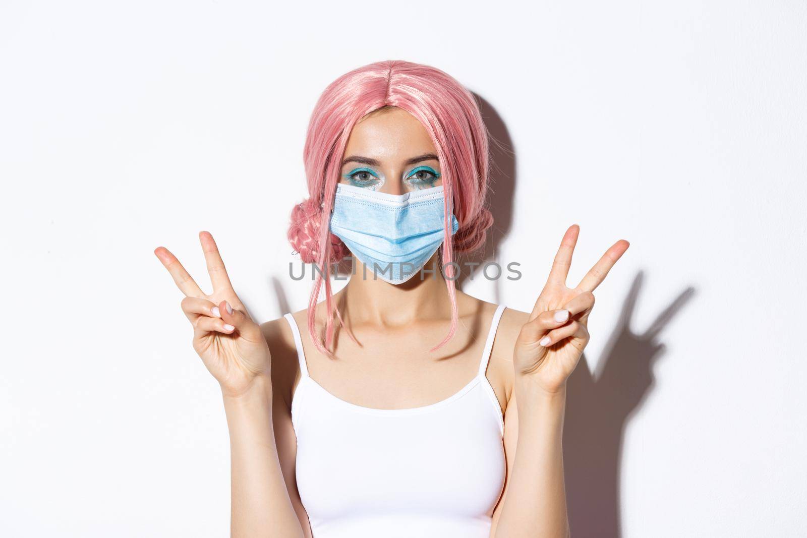 Coronavirus, social distancing and lifestyle concept. Close-up of pretty party girl in medical mask and pink wig, showing peace signs and smiling, standing over white background.