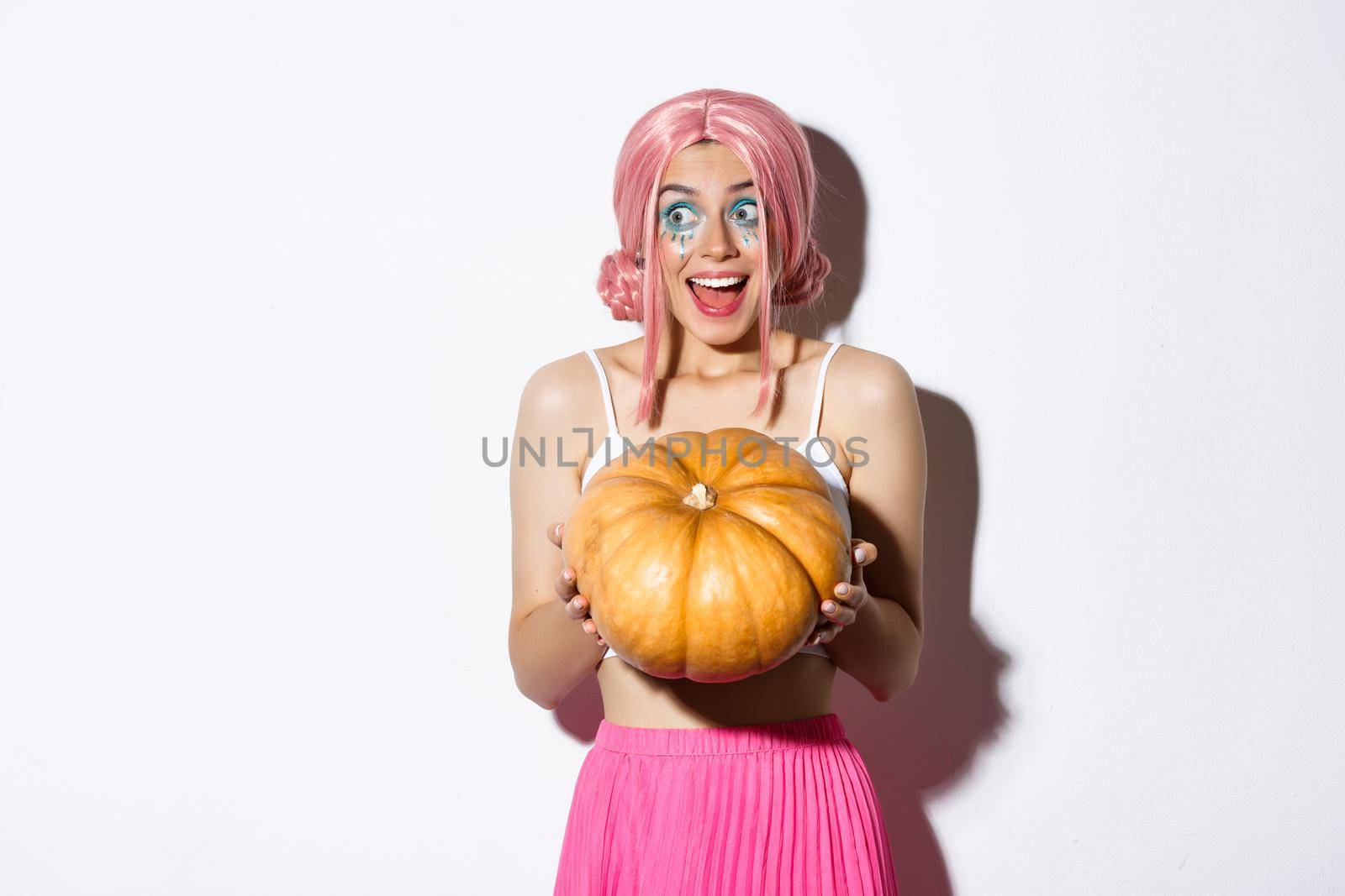 Image of excited attractive girl smiling, holding pumpkin for halloween, wearing pink wig for party and celebration, standing over white background.