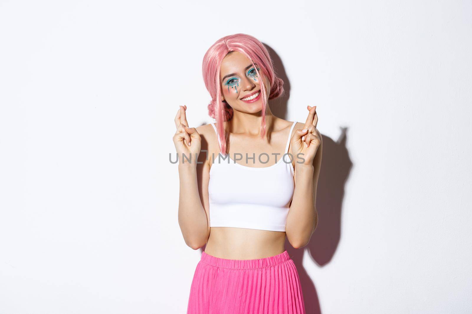 Portrait of lovely smiling woman in halloween costume, pink wig and bright makeup, looking hopeful at camera and making wish with fingers crossed.